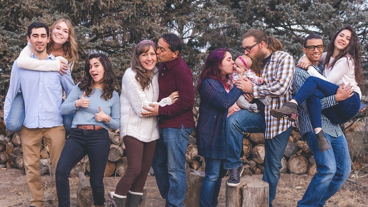 Foto em família, com uma mulher pulando nas costas de um homem, uma jovem com a boca aberta e os polegares para cima, um homem dando um beijo na bochecha de uma mulher, um homem e uma mulher beijando a cabeça de um bebê, e um homem segurando uma mulher no colo.