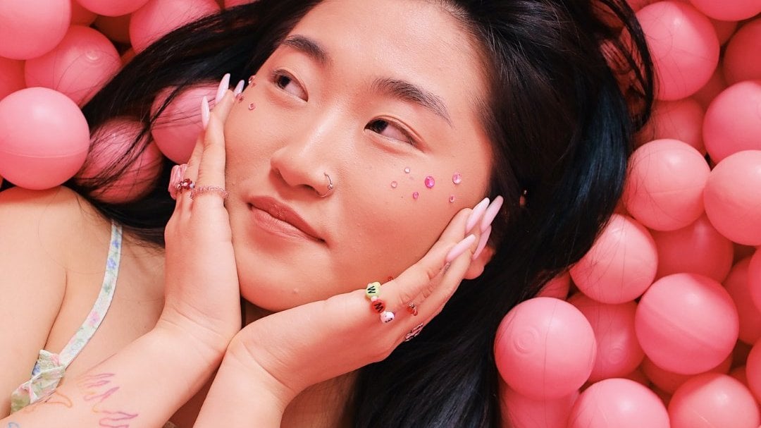 Menina asiática com as mãos em cada bochecha do rosto e olhando para o lado. Bolinhas rosas estão ao fundo