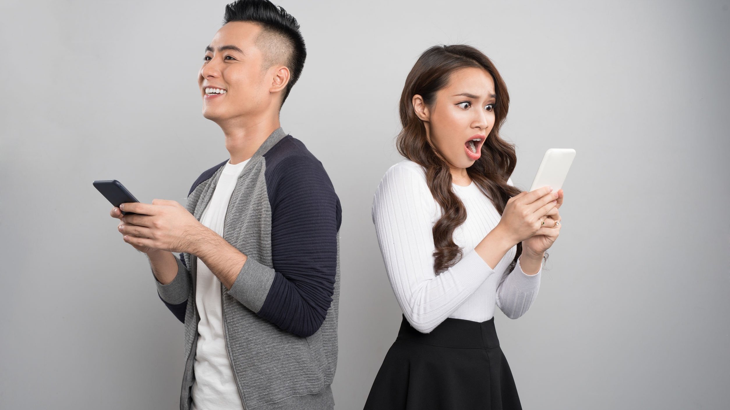 Homem asiática segurando celular com expressão sorridente, e mulher branca segurando celular com expressão surpresa.