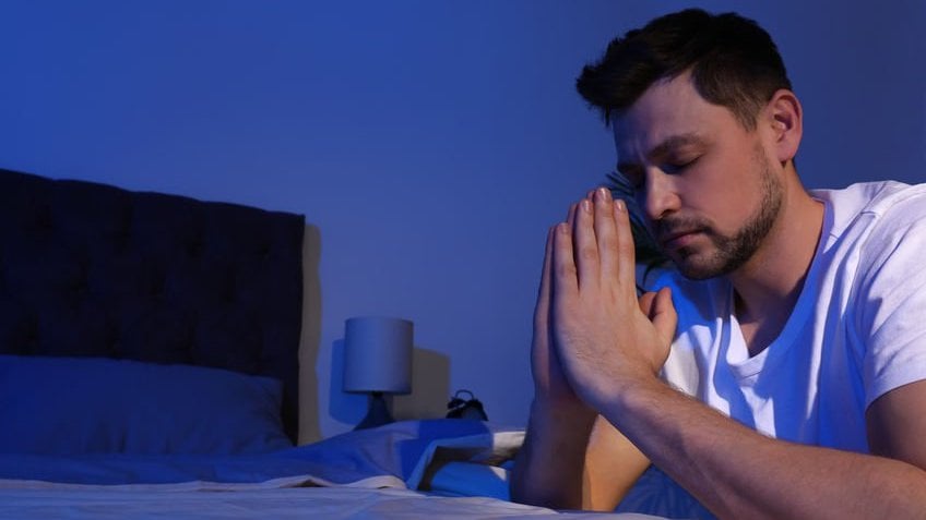 Homem ajoelhado à beira da cama em oração