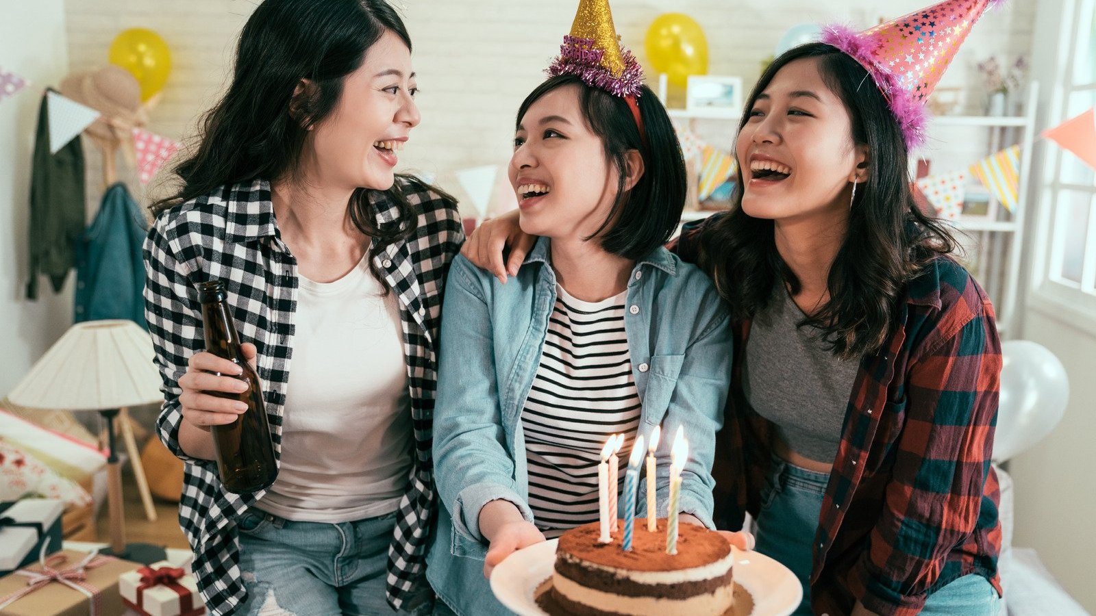 Três mulheres asiáticas com adereços comemorativos enquanto seguram bolo de aniversário.