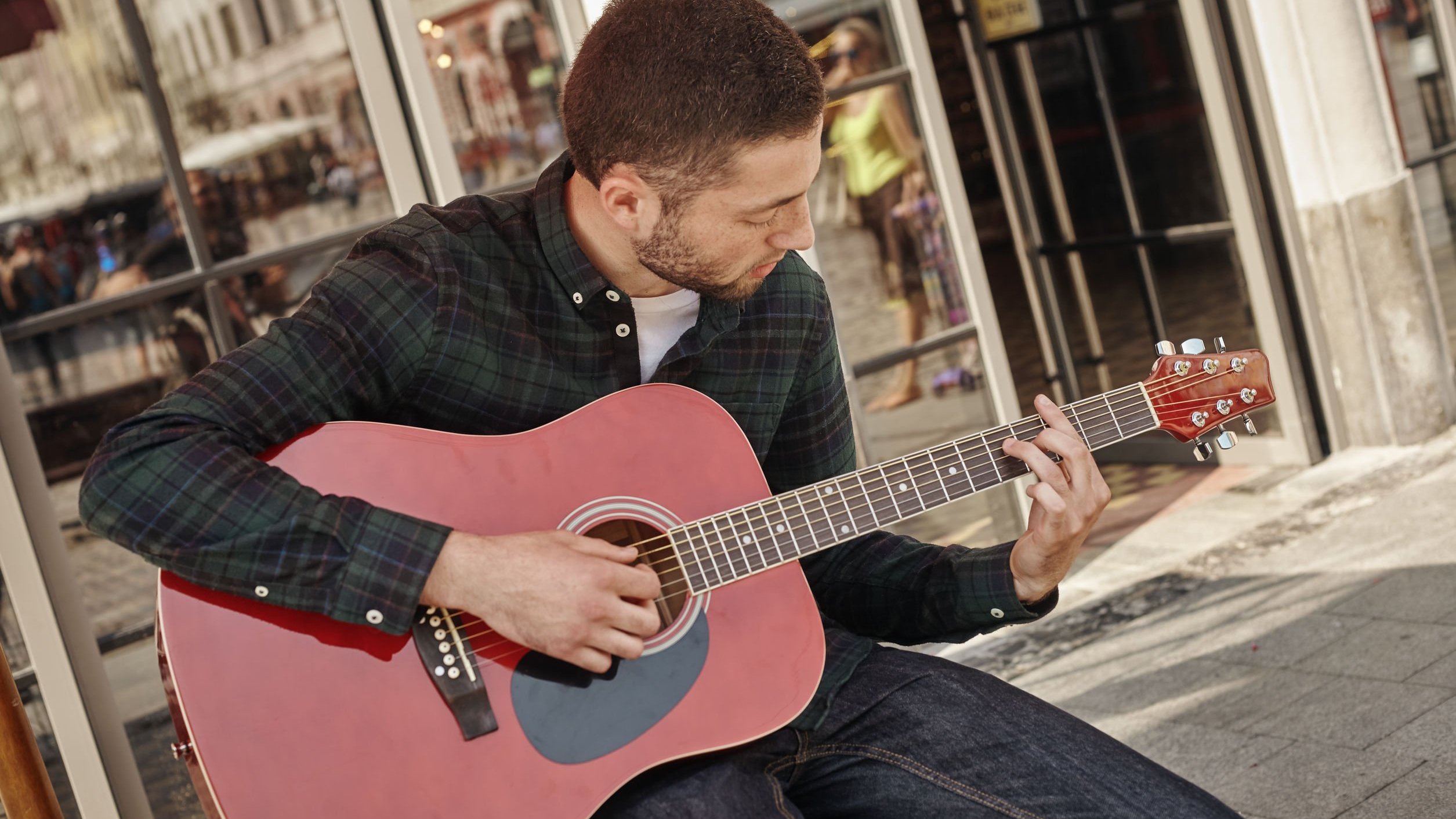 Homem branco sentado num banco tocando violão.