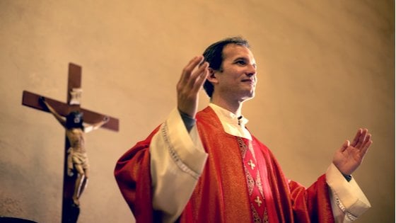 Padre com as mãos erguidas, sorrindo levemente. Atrás, uma cruz com a imagem de Jesus crucificado.