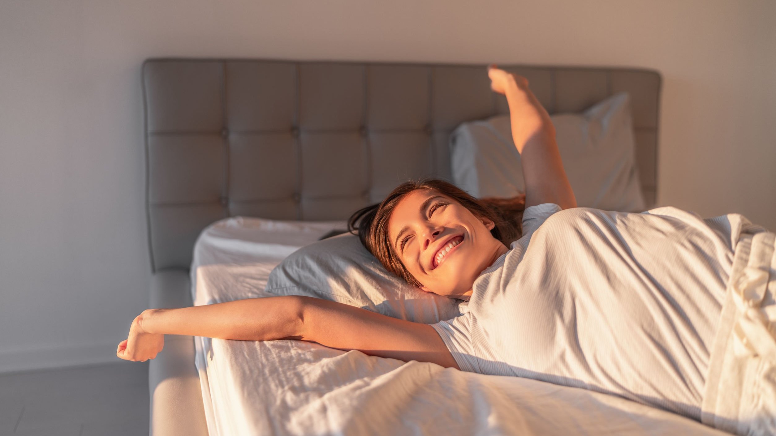 Deitada sobre uma cama, mulher estende os braços e sorri ao olhar para janela