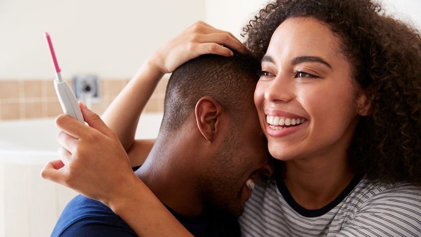 123rf. Mulher abraçando seu marido sorrindo com um teste de gravidez na mão.