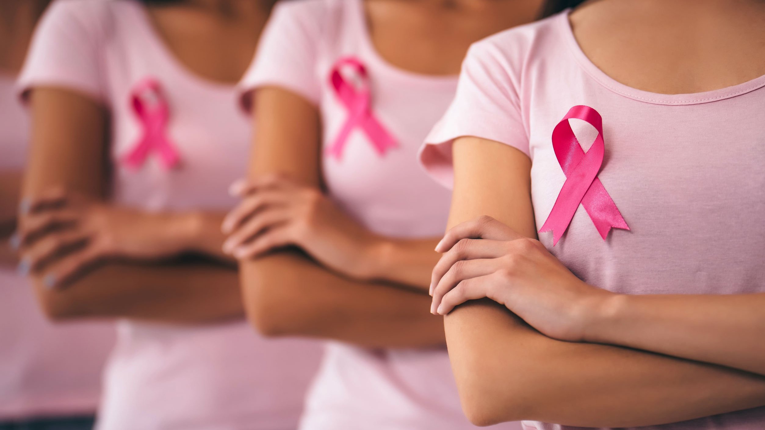 Mulheres em fila, de braços cruzados, com fitas de conscientização do câncer de mama na camiseta.