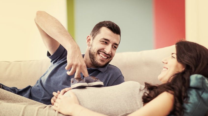 Homem sorridente segurando dois sapatinhos de bebê na barriga de mulher gestante.