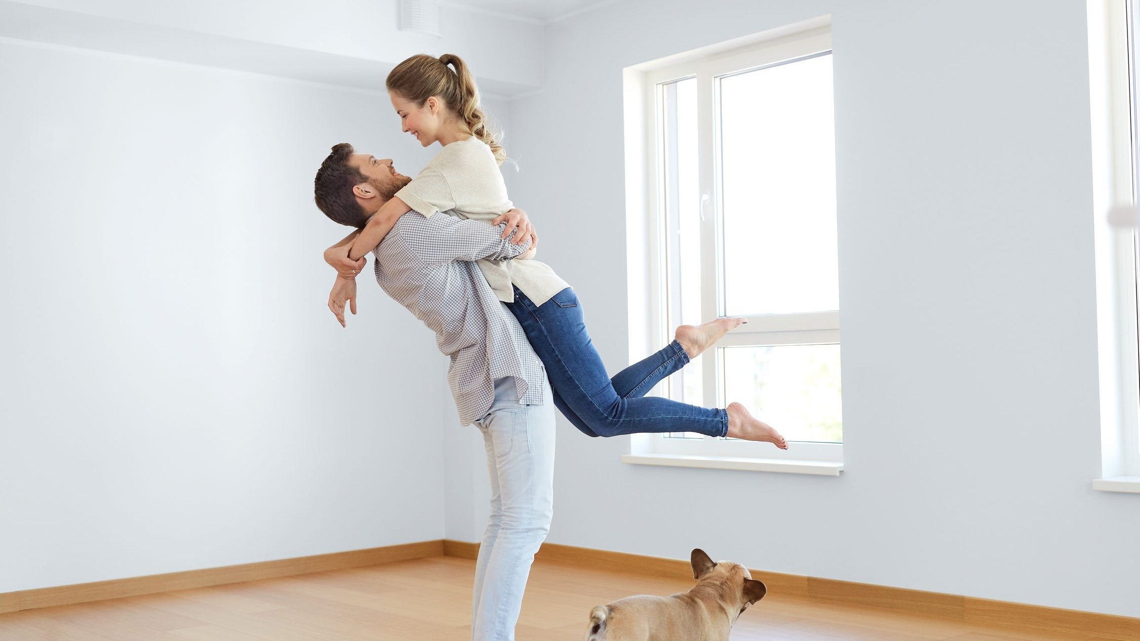 Homem levantando mulher enquanto a abraça, em um cômodo vazio, com piso de madeira e paredes brancas. Um cachorro no chão olha a cena.