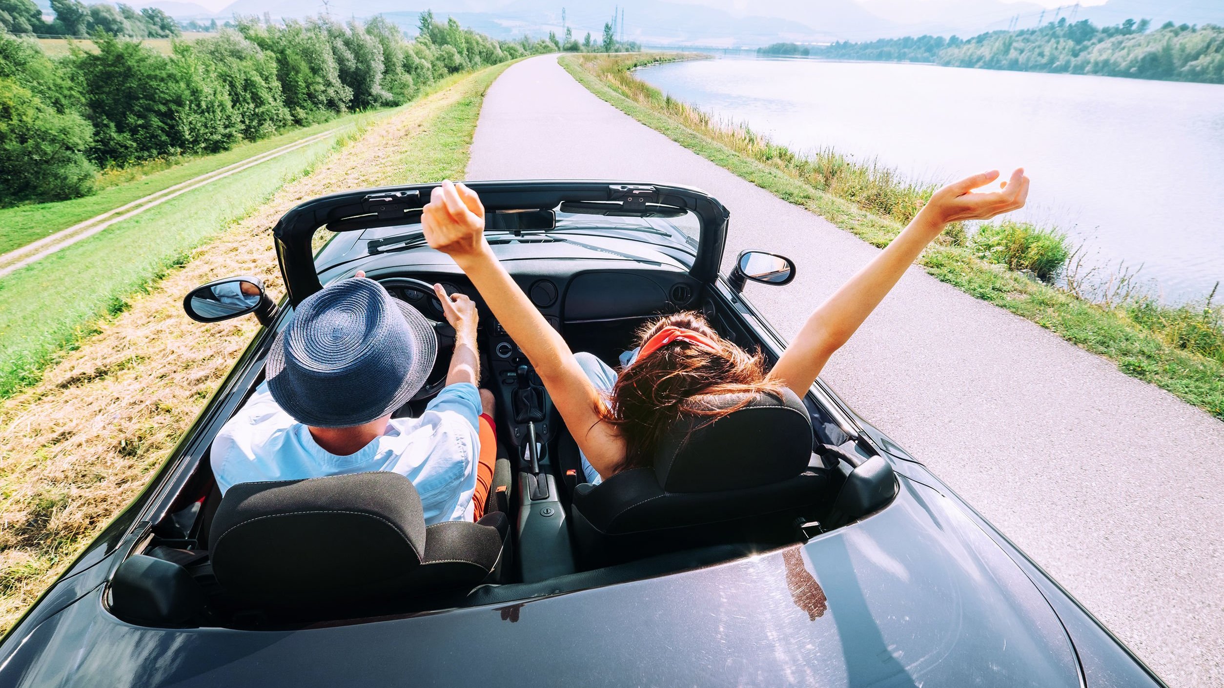 Homem e mulher em carro conversível. O homem dirige e a mulher está com os braços para o alto.