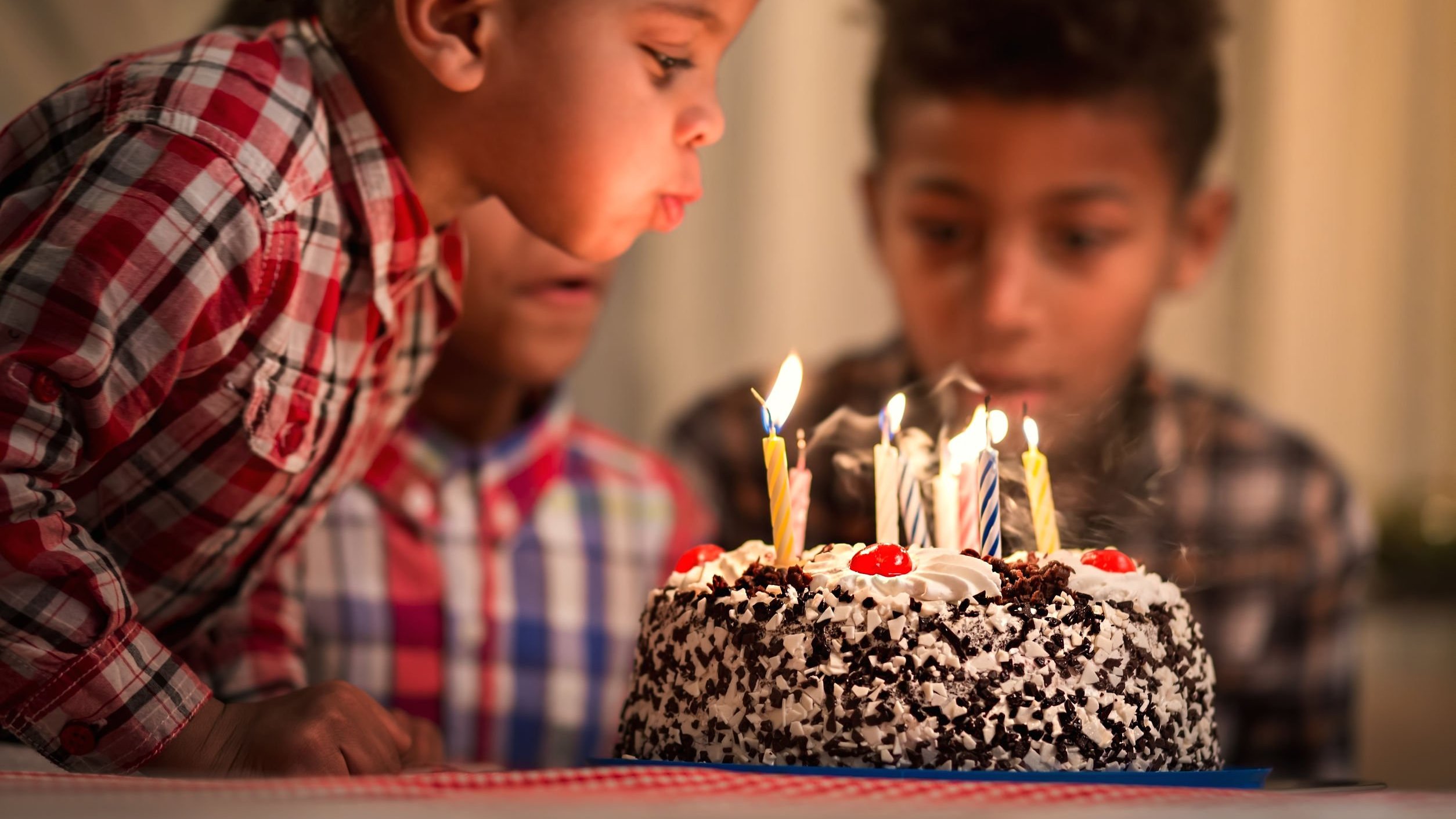 Menino pequeno assoprando velas em cima de um bolo, e um menino mais velho ao seu lado olha a cena.