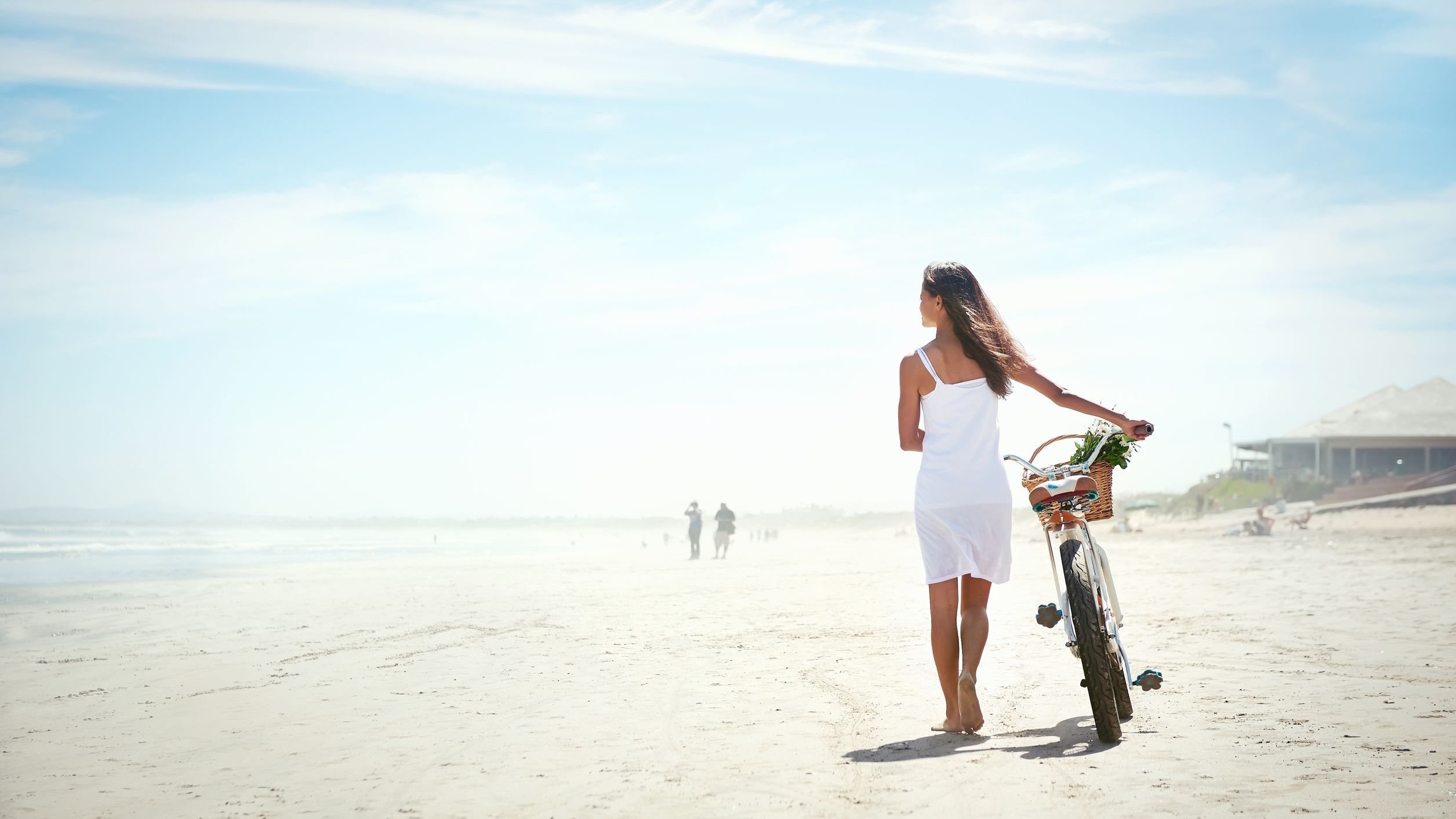 Mulher caminhando descalça na praia, guiando sua bicicleta com as mãos. Ela olha para o amor, e o céu está azul.
