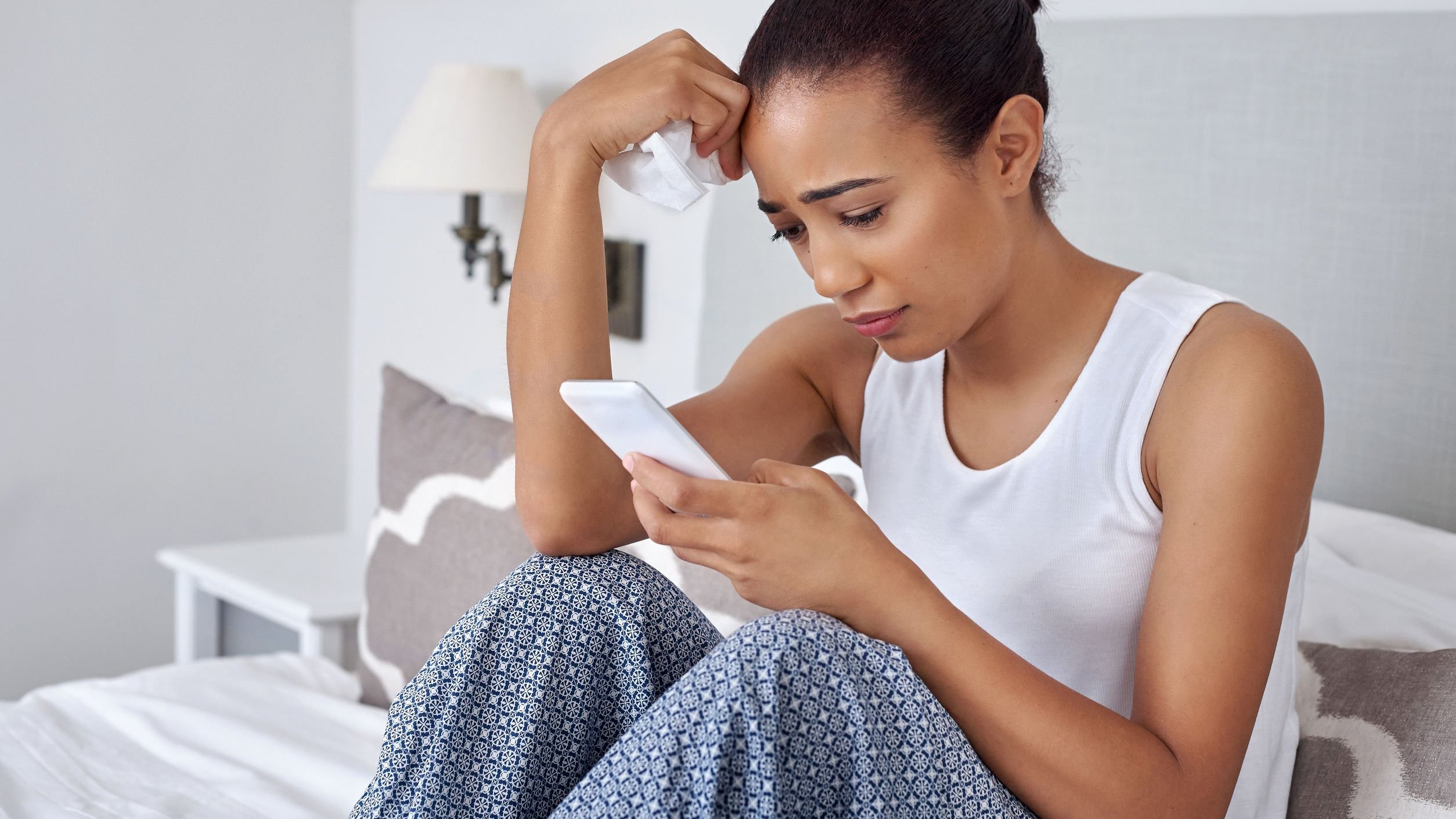 Mulher triste olhando celular enquanto segura um lenço de papel com a outra mão.