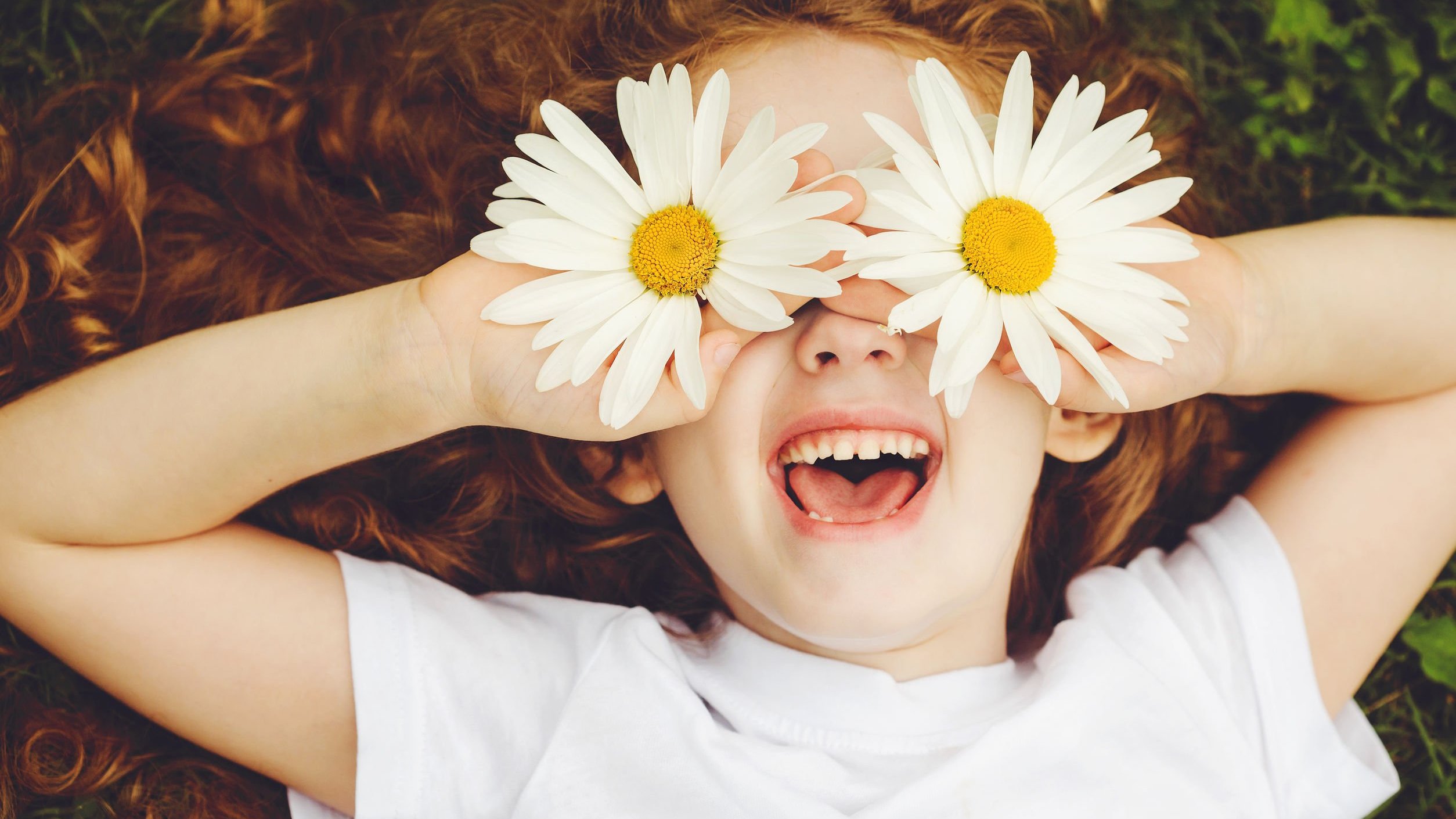 Menina branca de cabelos ruivos deitada na grama, com flores amarelas levadas aos olhos.