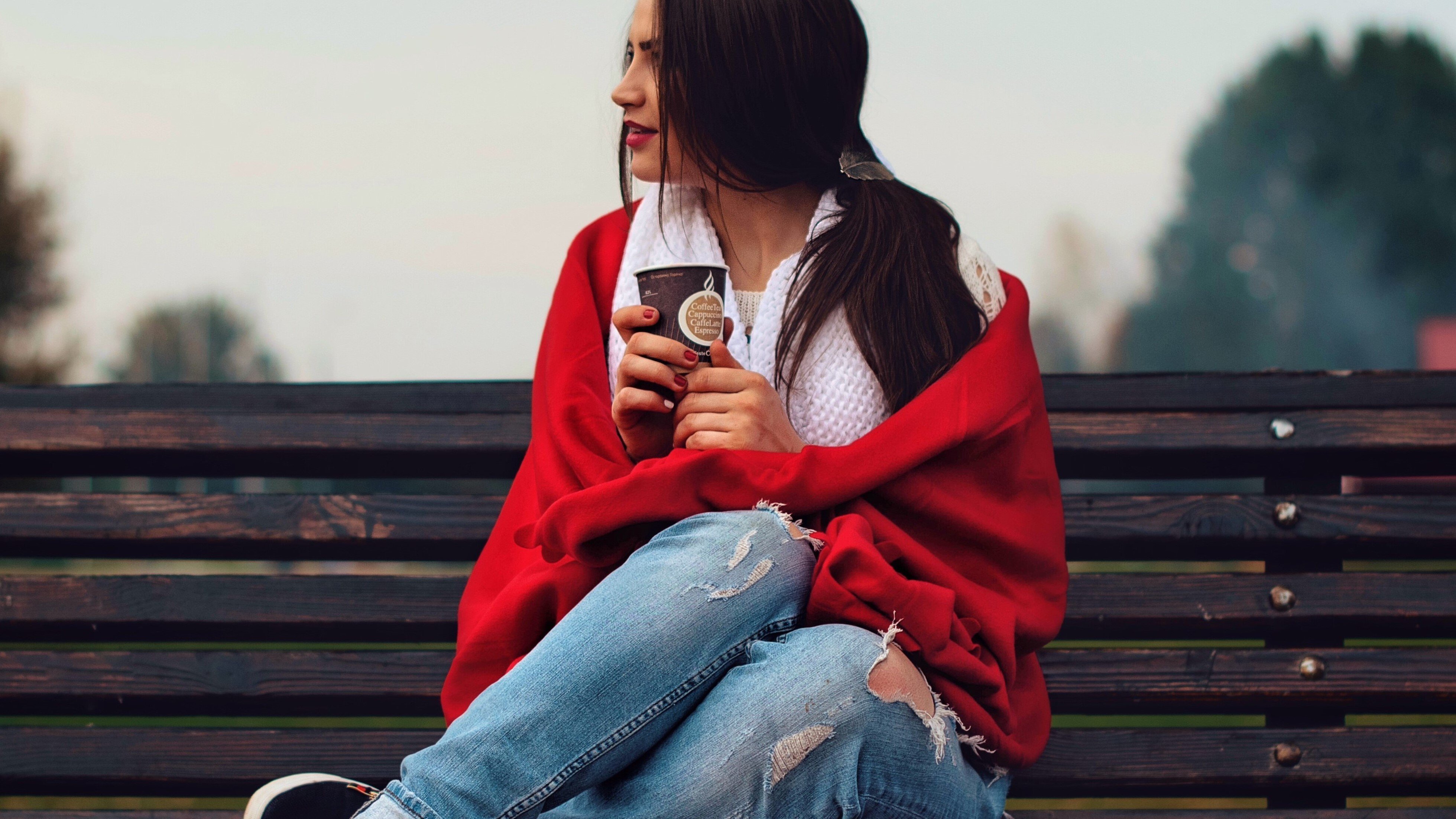 Mulher sentada em um banco de madeira olhando para o lado pensativa segurando um copo de café