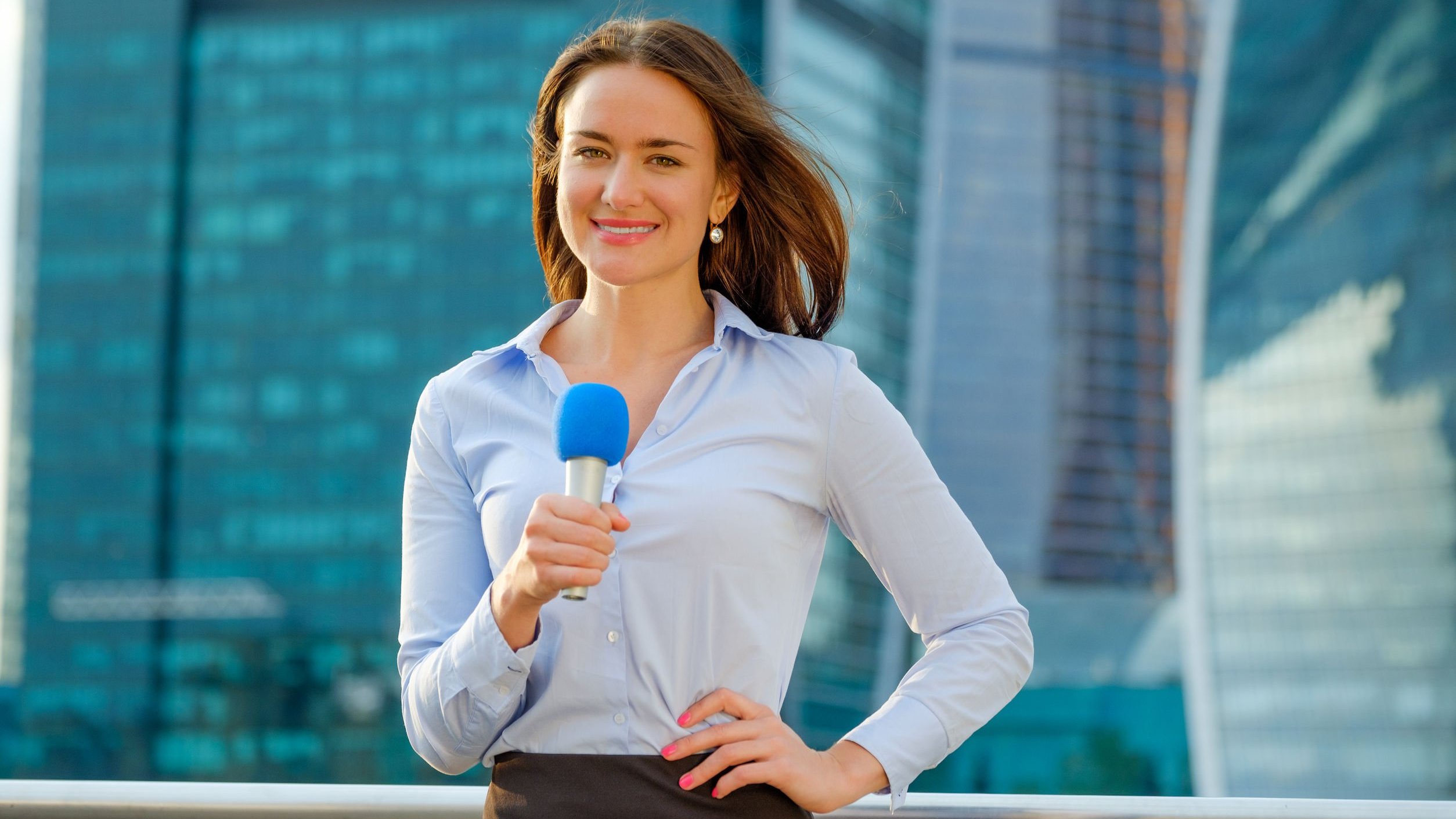 Mulher com camisa social azul, mão esquerda apoiada na cintura e segurando um microfone com a mão direita. Ao fundo dela, vemos prédios comerciais com fachadas espelhadas.