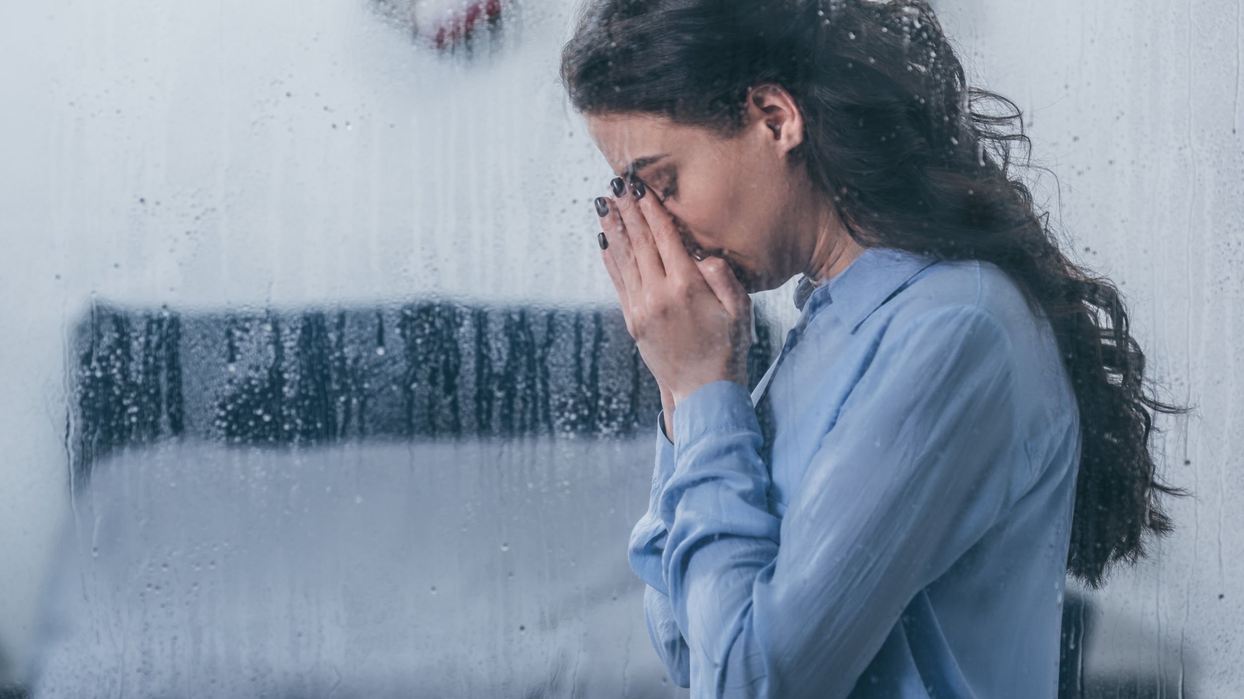 Mulher rezando ao aldo de uma janela molhada pela chuva