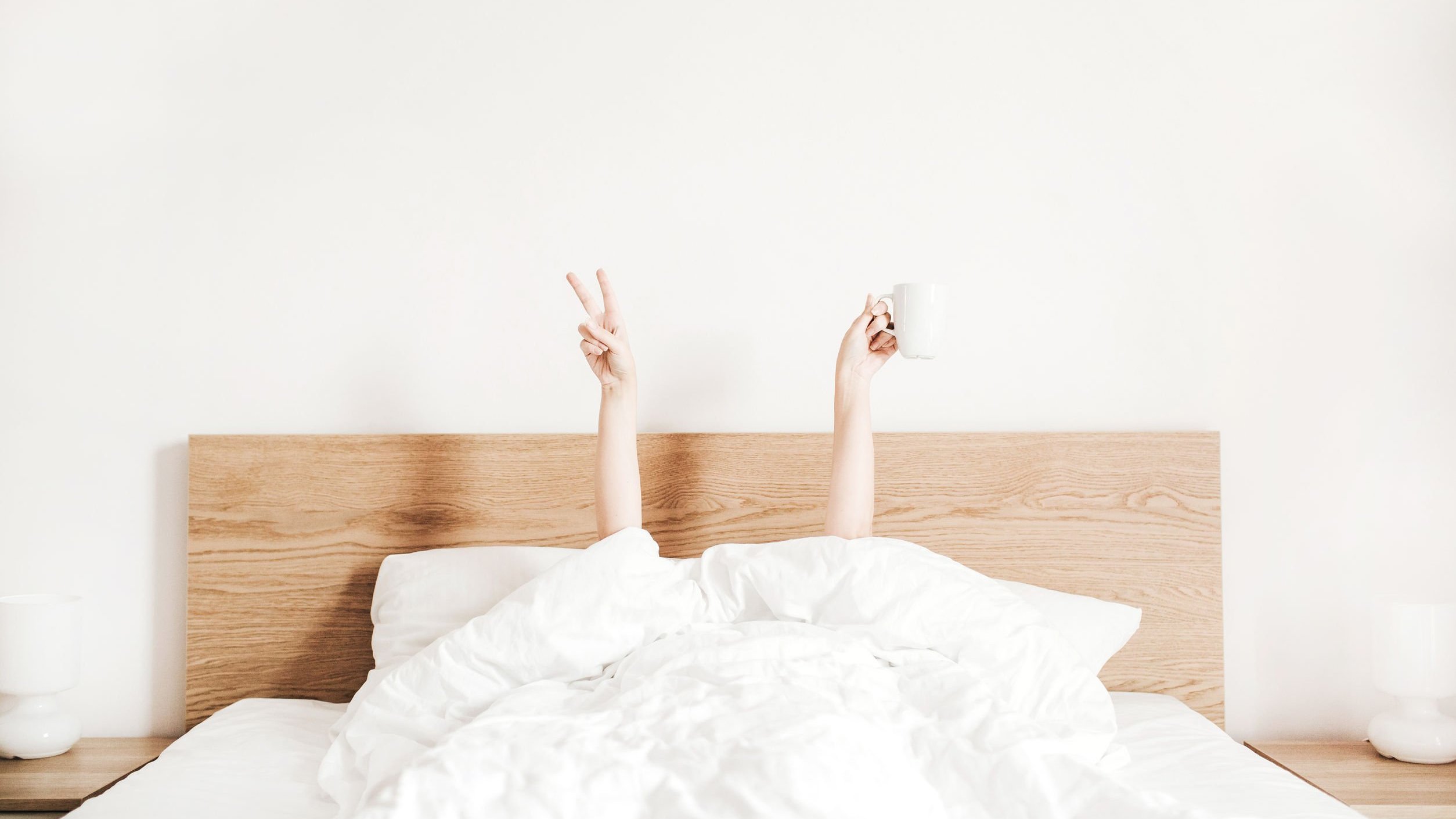 Pessoa deitada na cama apenas com as mãos levantadas: uma fazendo o sinal da paz e a outra segurando uma xícara de café.