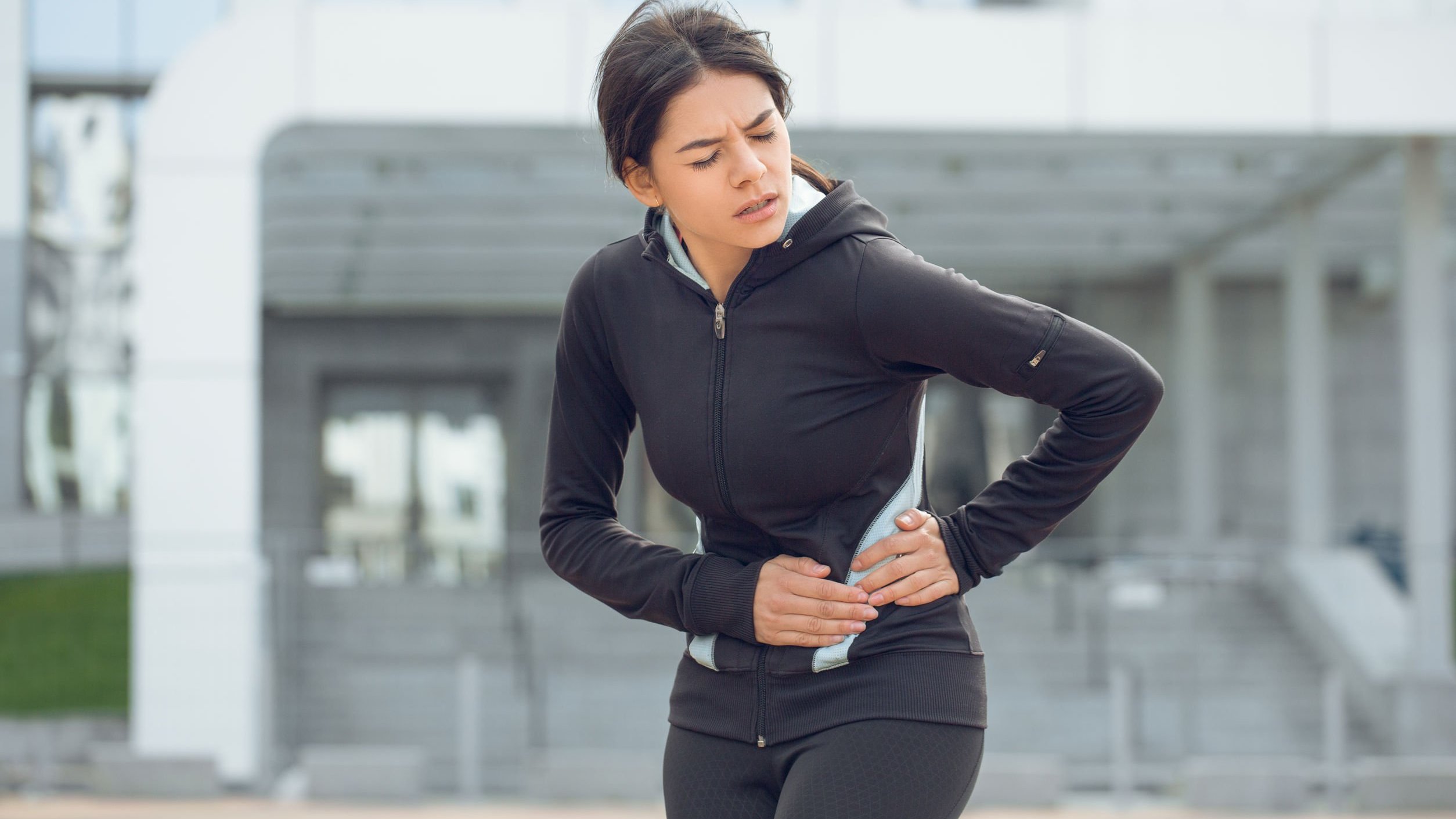 Mulher com roupa de ginástica, com os olhos fechados e as duas mãos na cintura como se estivesse com dor.