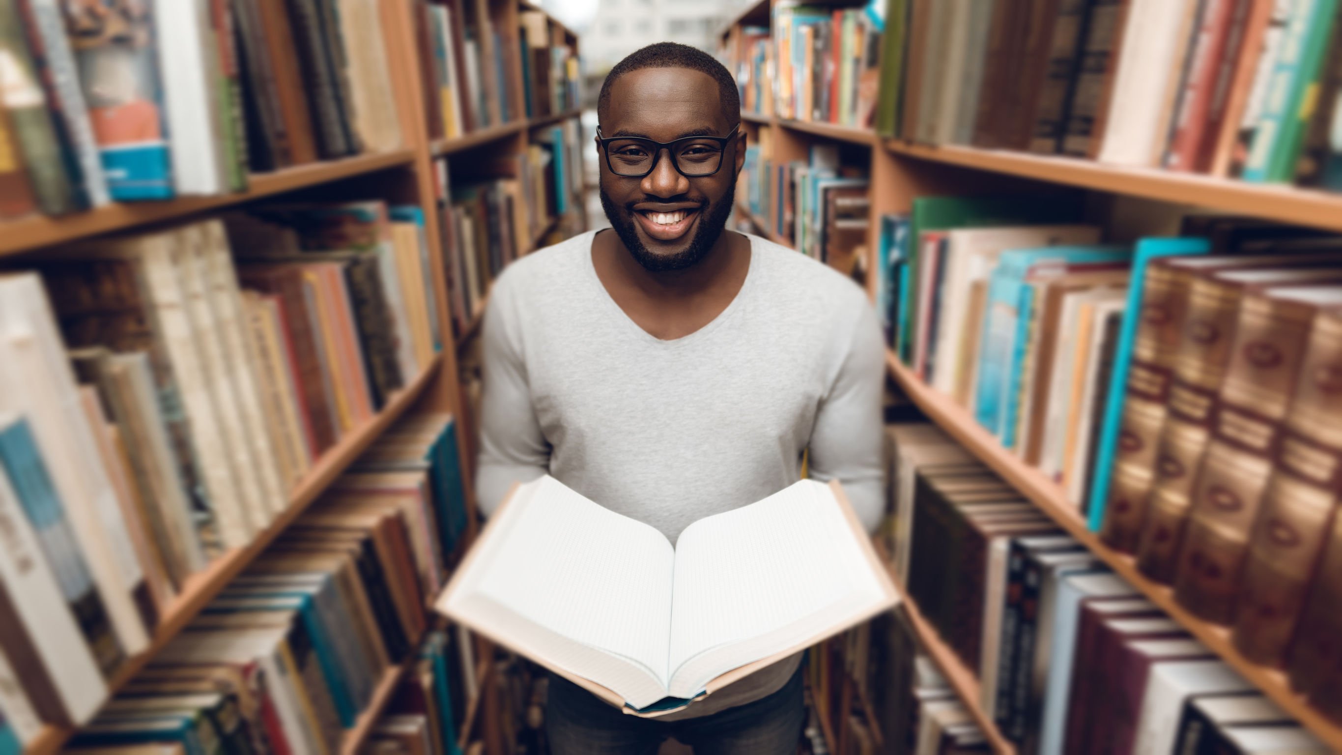 Homem segurando um livro aberto entre prateleiras de biblioteca.