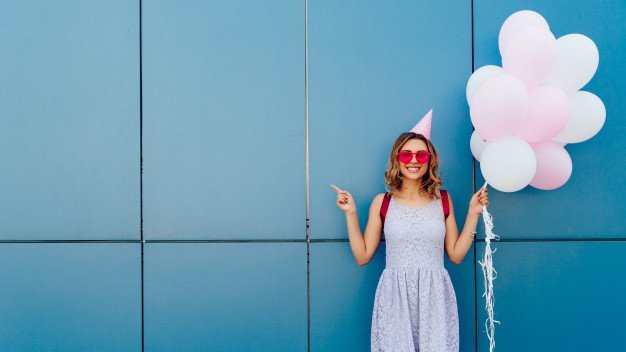 Freepik Garota encostada em uma parede verde, com chapéu de aniversário e segurando balões.