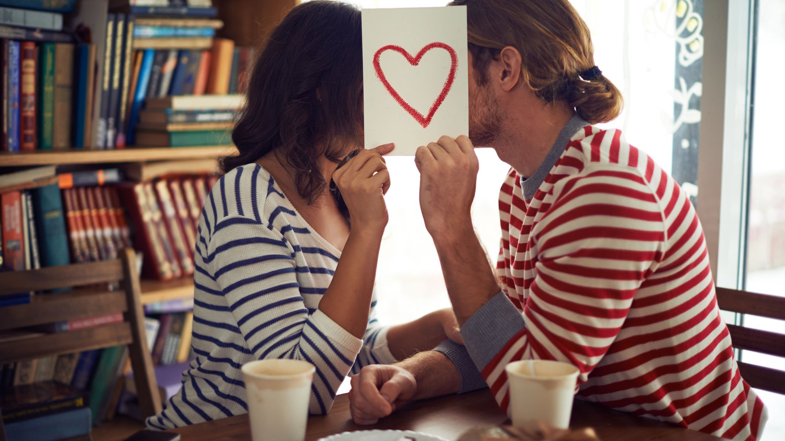 Foto de casal se escondendo atrás de cartão com desenho de coração