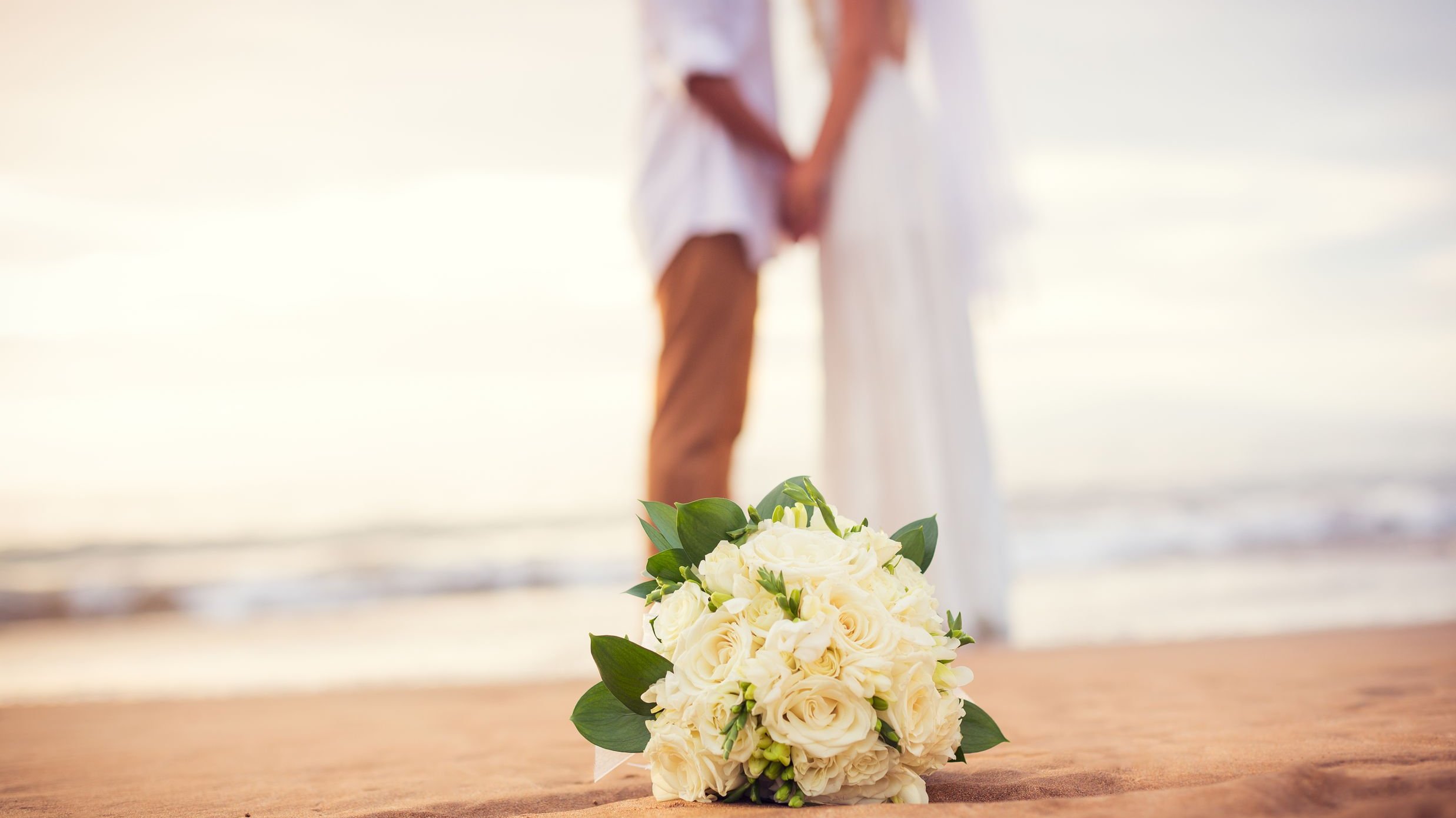 Buquê de flores no chão da praia, ao fundo, desfocado, há um casal de noivos vestidos de branco