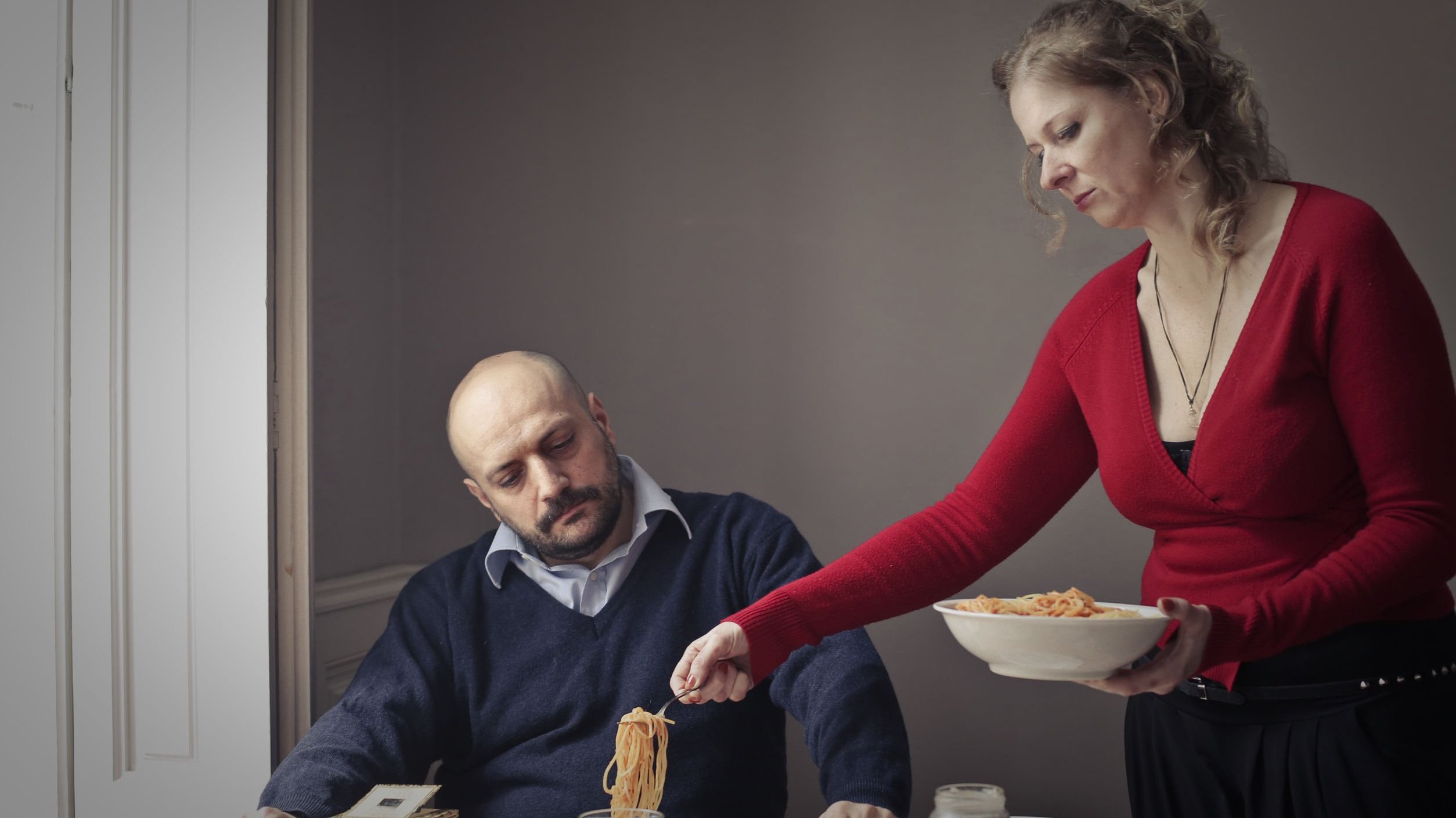 Homem sentado em mesa de jantar com mulher lhe servindo uma porção de macarrão. O homem parece impaciente e a mulher tem uma expressão cansada.