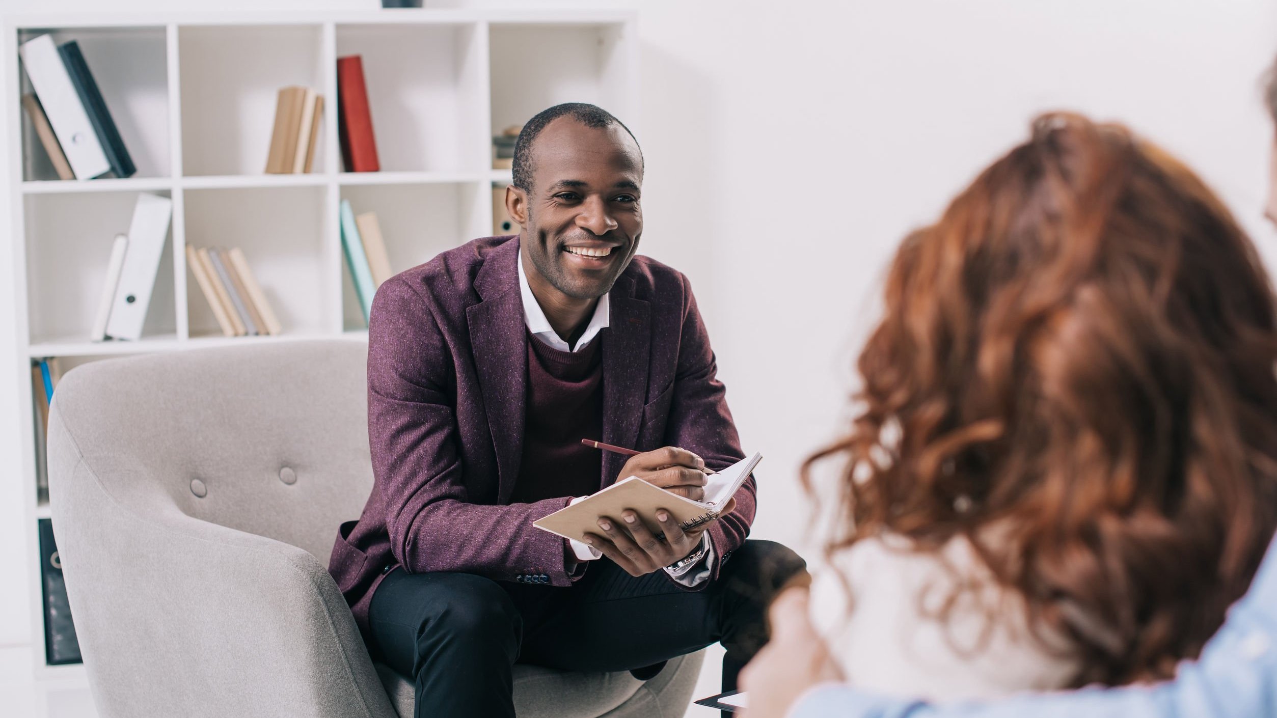 Terapeuta, homem negro, sentado sorridente conversando com mulher e fazendo anotações