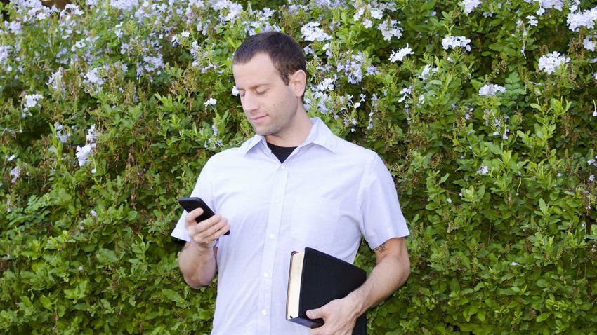 Homem com celular na mão sorrindo e bíblia embaixo do braço