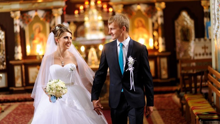 Mulher com vestido de noiva e homem com terno e gravata saindo de igreja de mãos dadas após seu casamento.