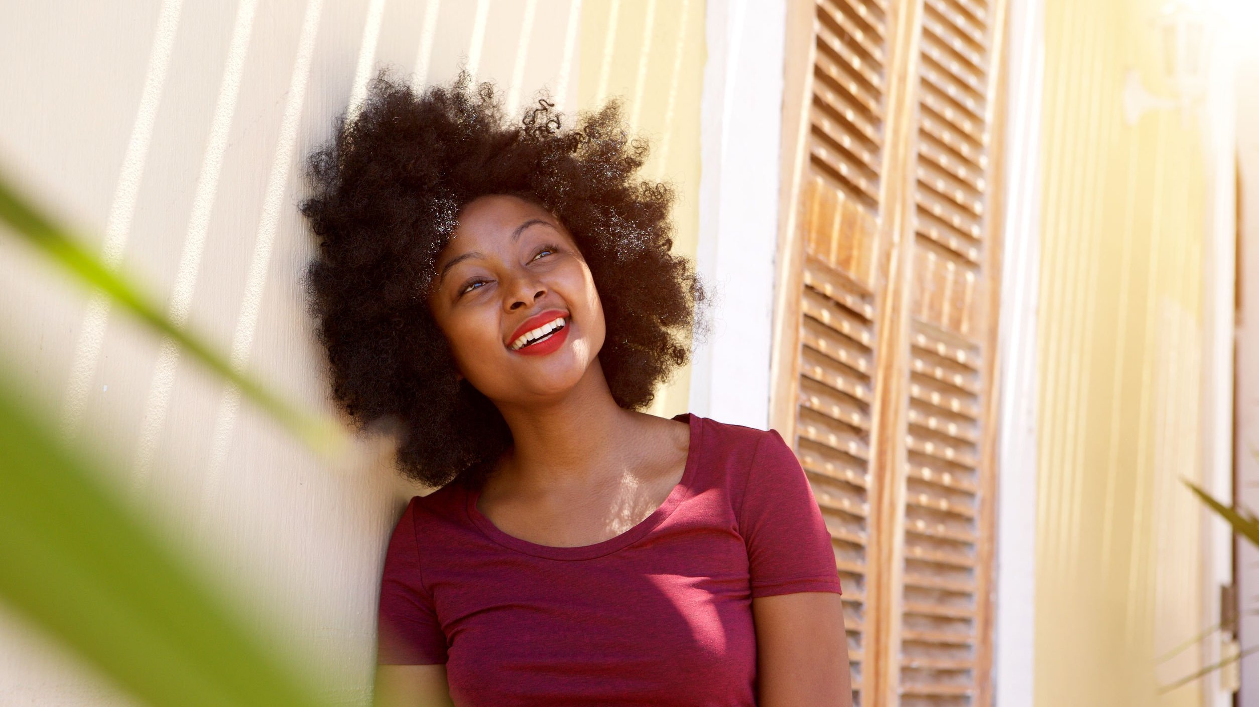 Mulher sorridente apoiada na parede, em pé, do lado de fora de uma casa.