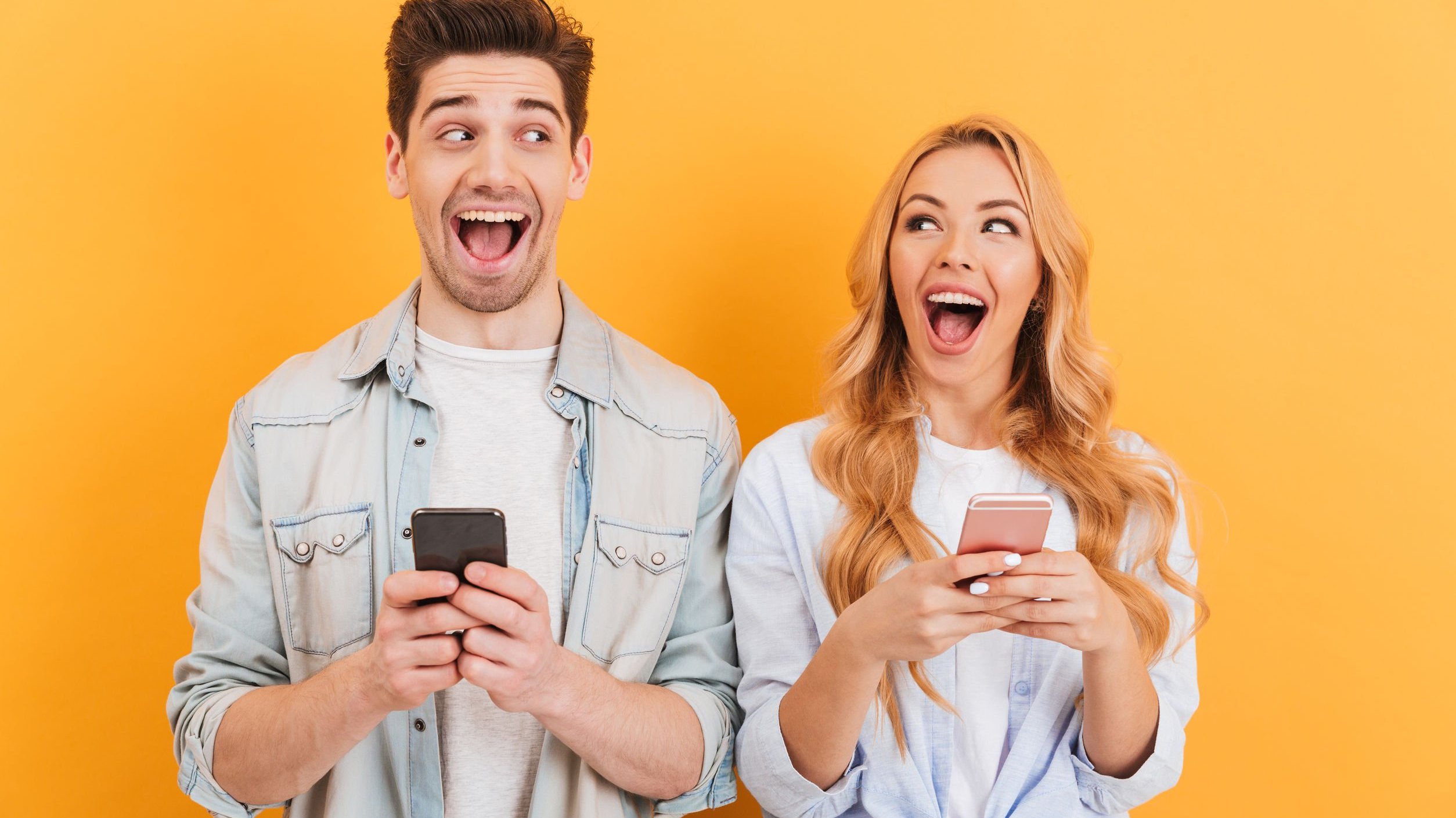 Homem e mulher lado a lado, rindo enquanto seguram seus smarthphones e olham um para o outro.