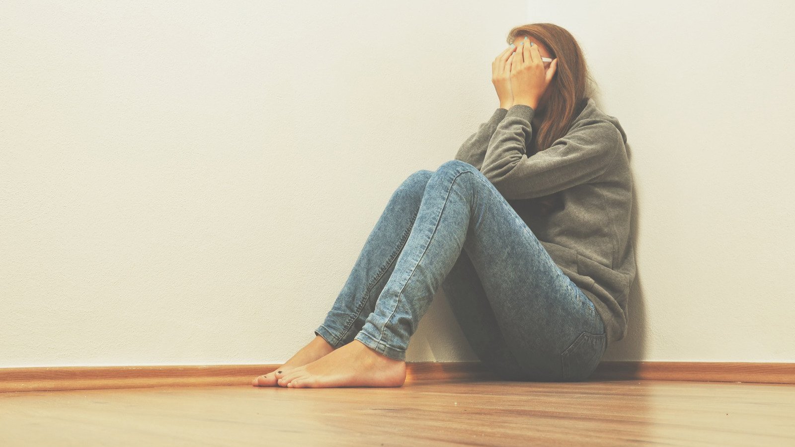nixki / Canva PRO Mulher branca sentada no chão com braços encolhidos e cabeça entre mãos.