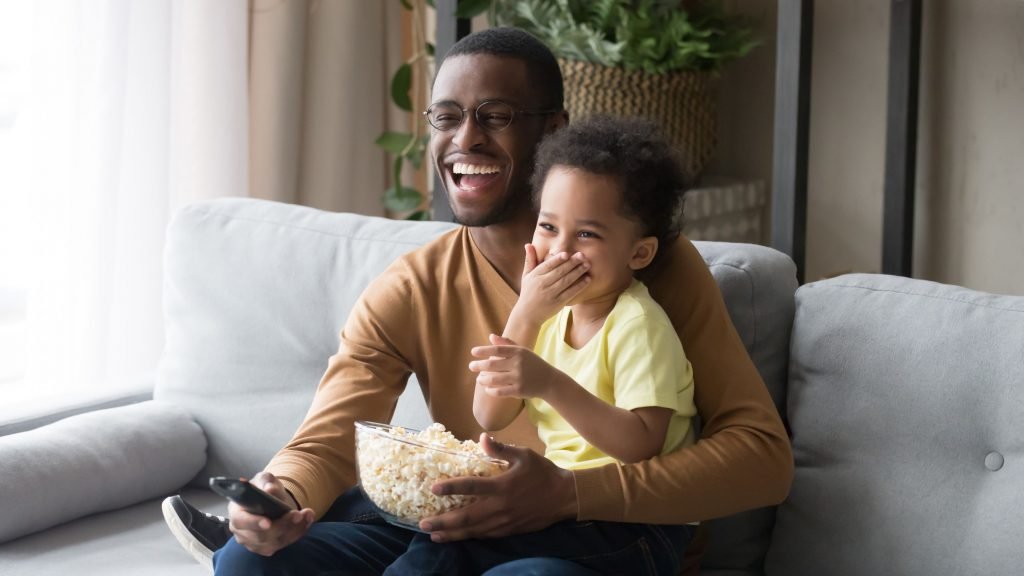 123rf Pai e filha sentados no sofá comendo pipoca e assistindo televisão.