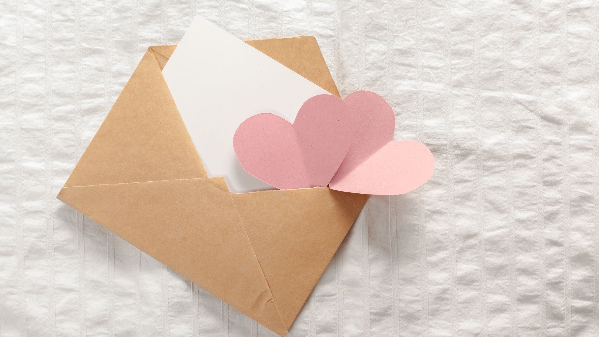 Envelope marrom aberto com carta branca e corações rosa claro saindo de dentro dele.