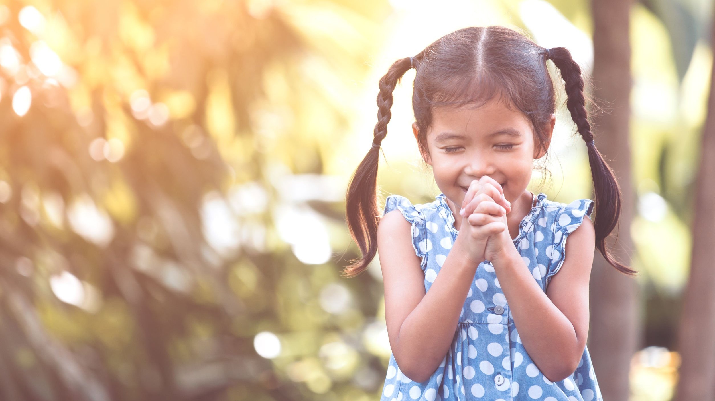 Menina com as mãos unidas e olhos fechados, orando, enquanto toma sol