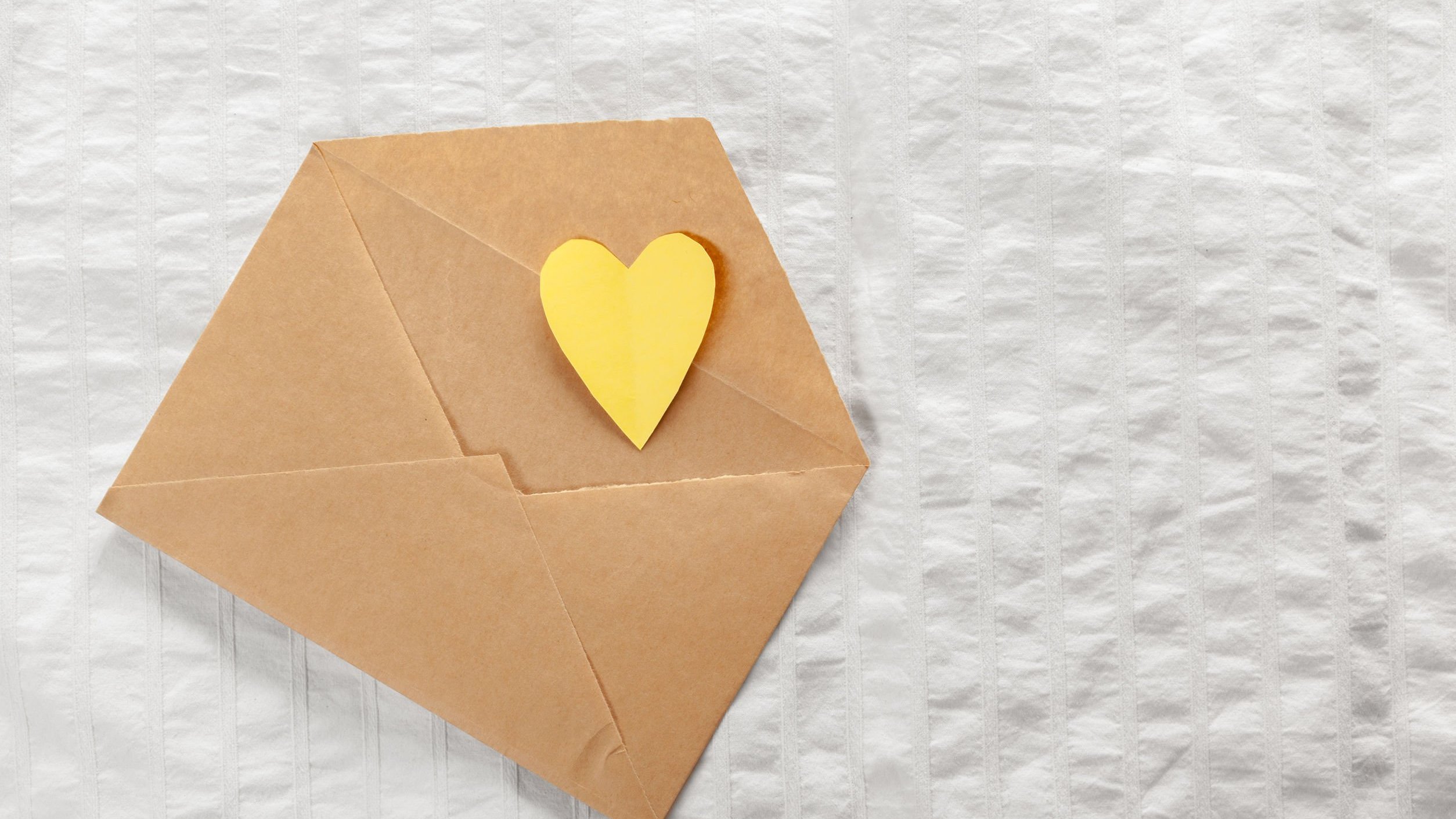 Envelope marrom aberto, com coração amarelo saindo dele, colocado sob mesa branca