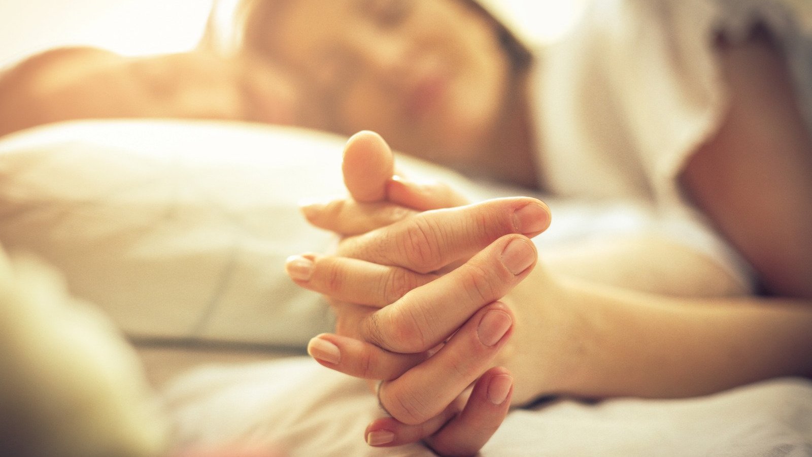 Homem e mulher brancos de mãos dadas, deitados numa cama.