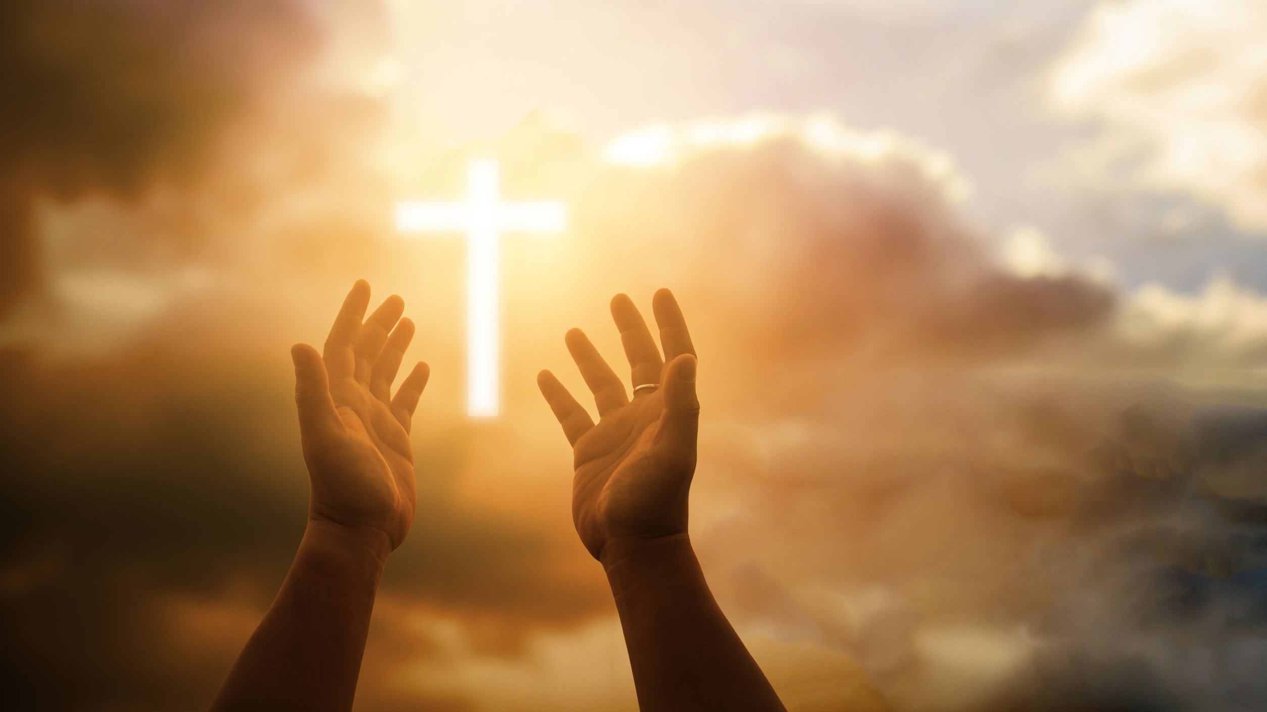 Mãos levantadas em direção ao céu, com uma luz em forma de cruz em meio as nuvens