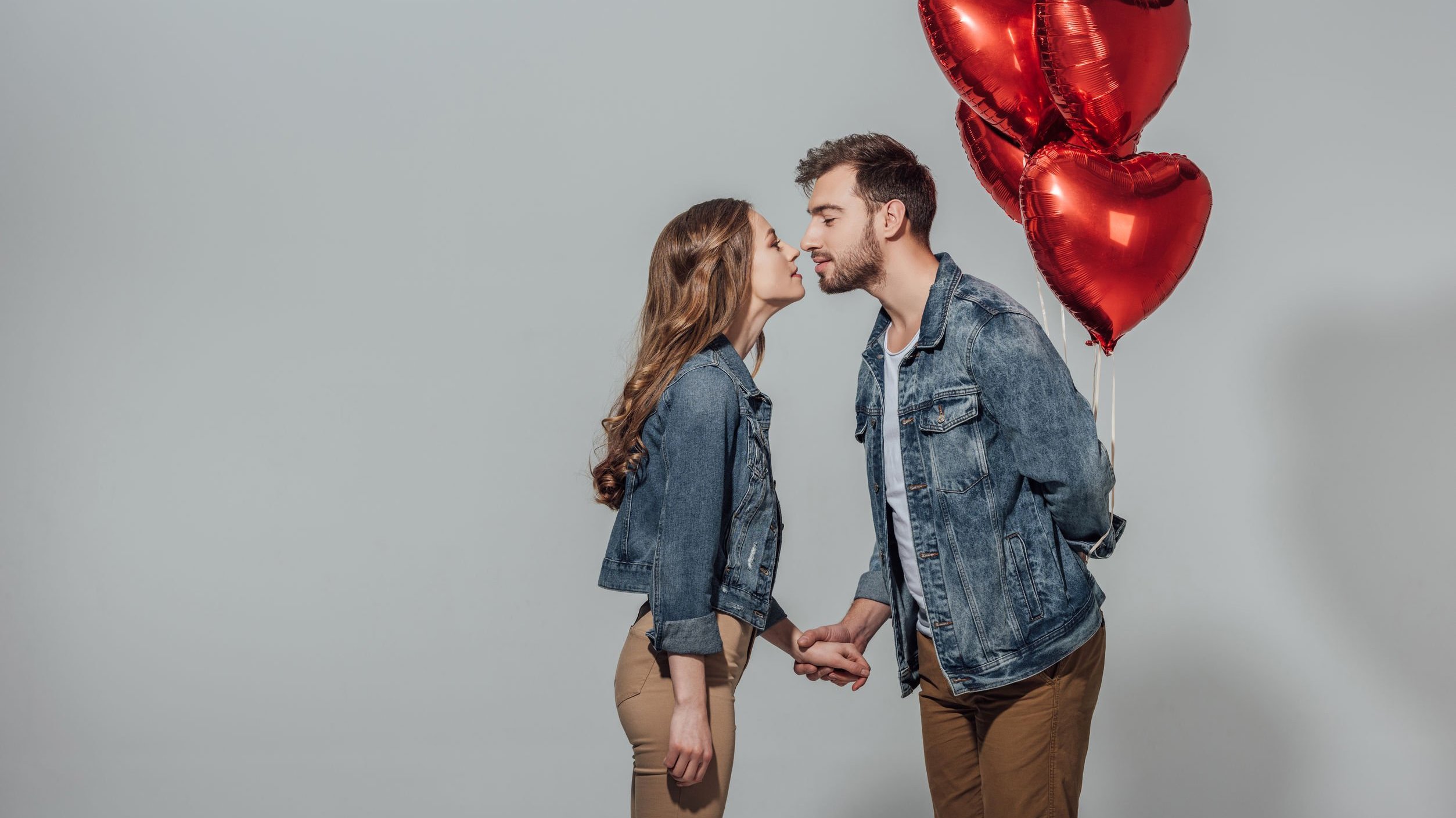 Homem segurando balão de corações com mão atrás das costas e beijando mulher