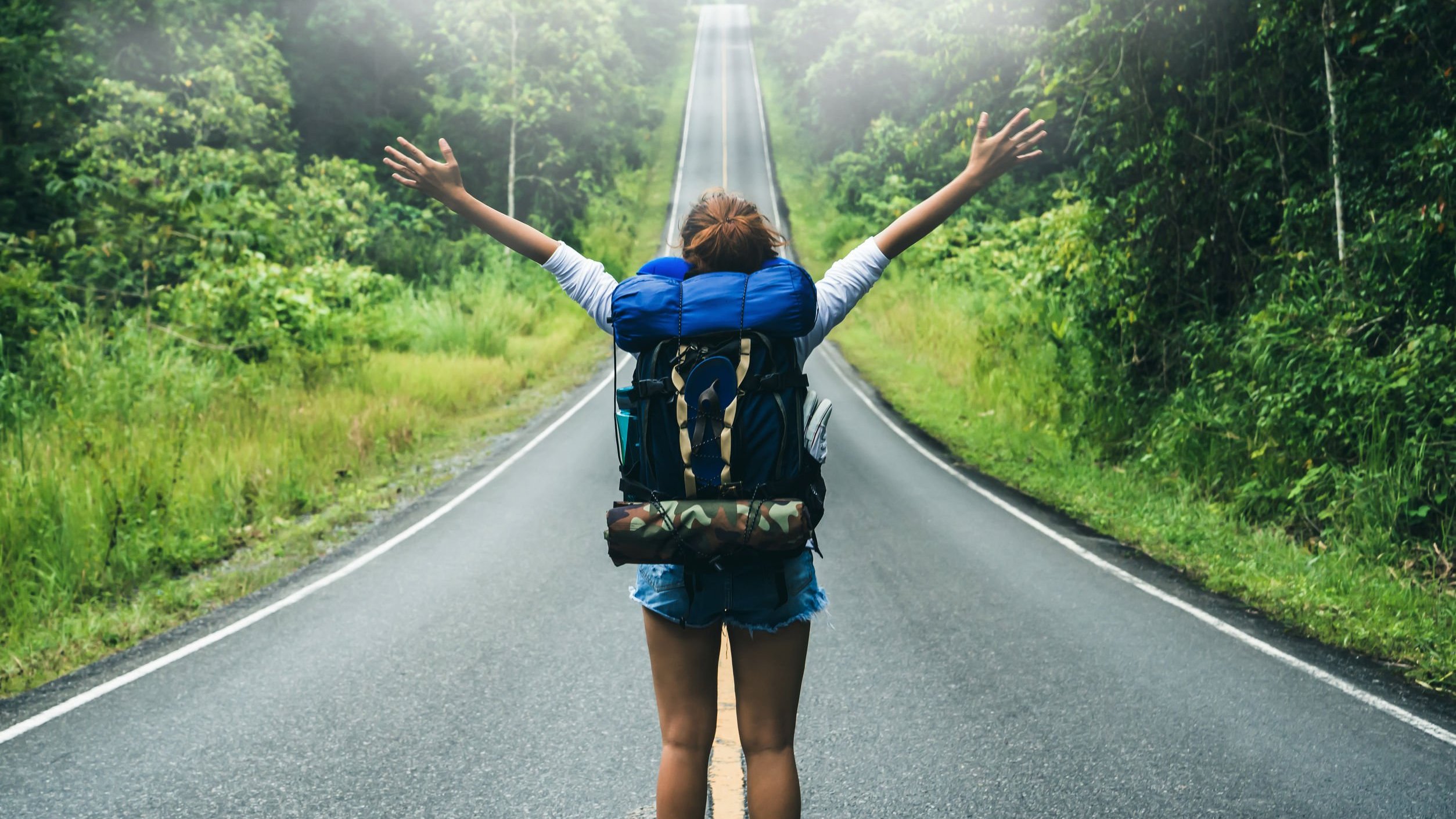 Mulher usando mochila de camping, com braços abertos olhando para estrada cercada de floresta