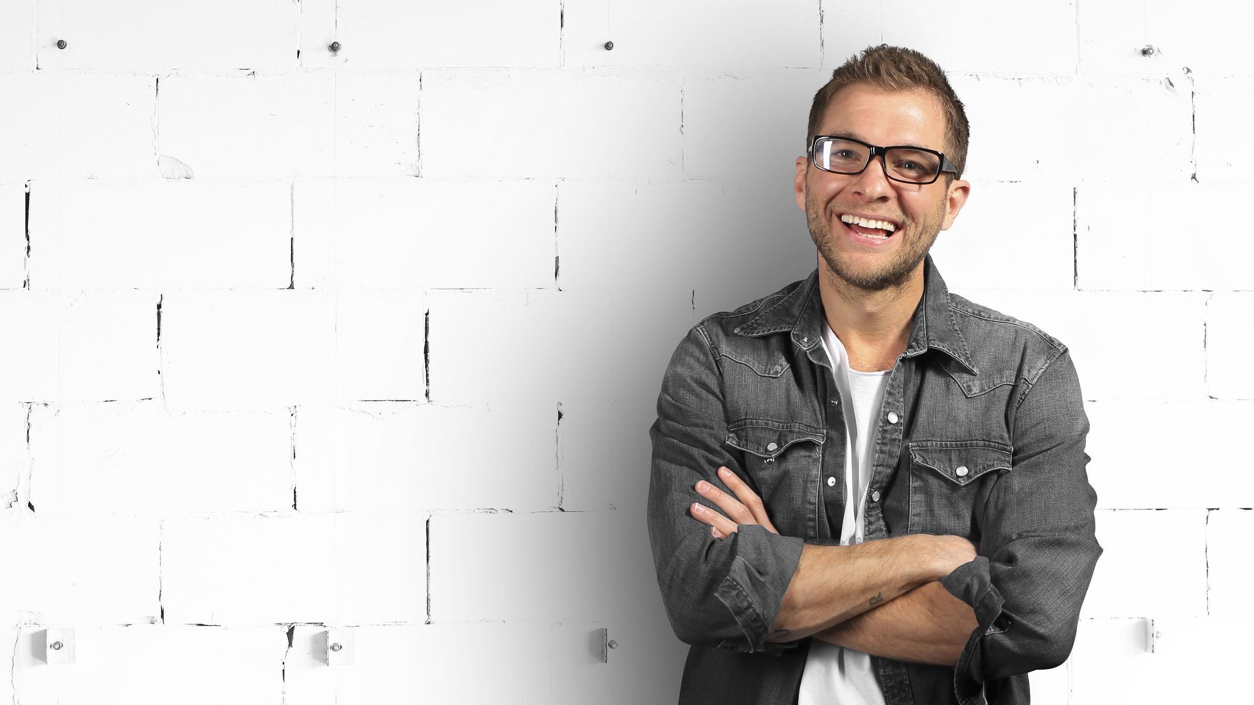 Homem sorrindo de braços cruzados encostado em parede de tijolos brancos