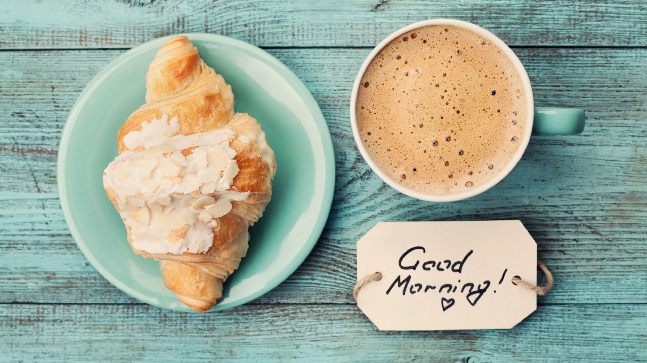 Foto de um croissant com uma xícara de café com leite. Ao lado, uma plaquinha com o escrito 'good morning' (bom dia em inglês'