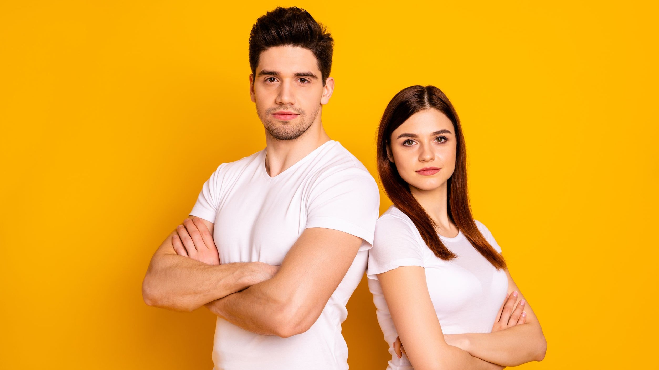 Homem e mulher posando de braços cruzados lado a lado, em frente de fundo amarelo