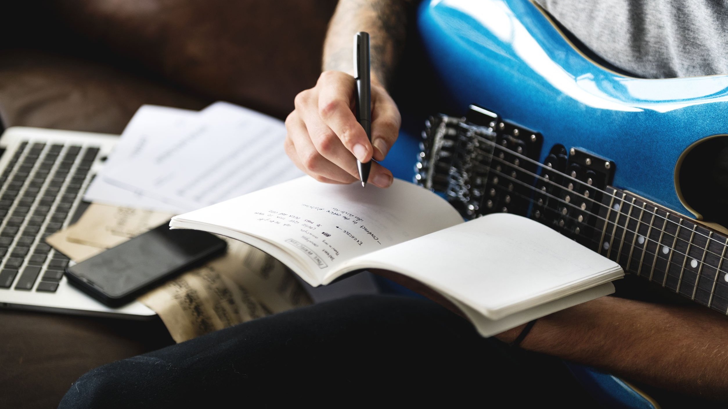 Homem sentado com guitarra no colo escrevendo em caderno