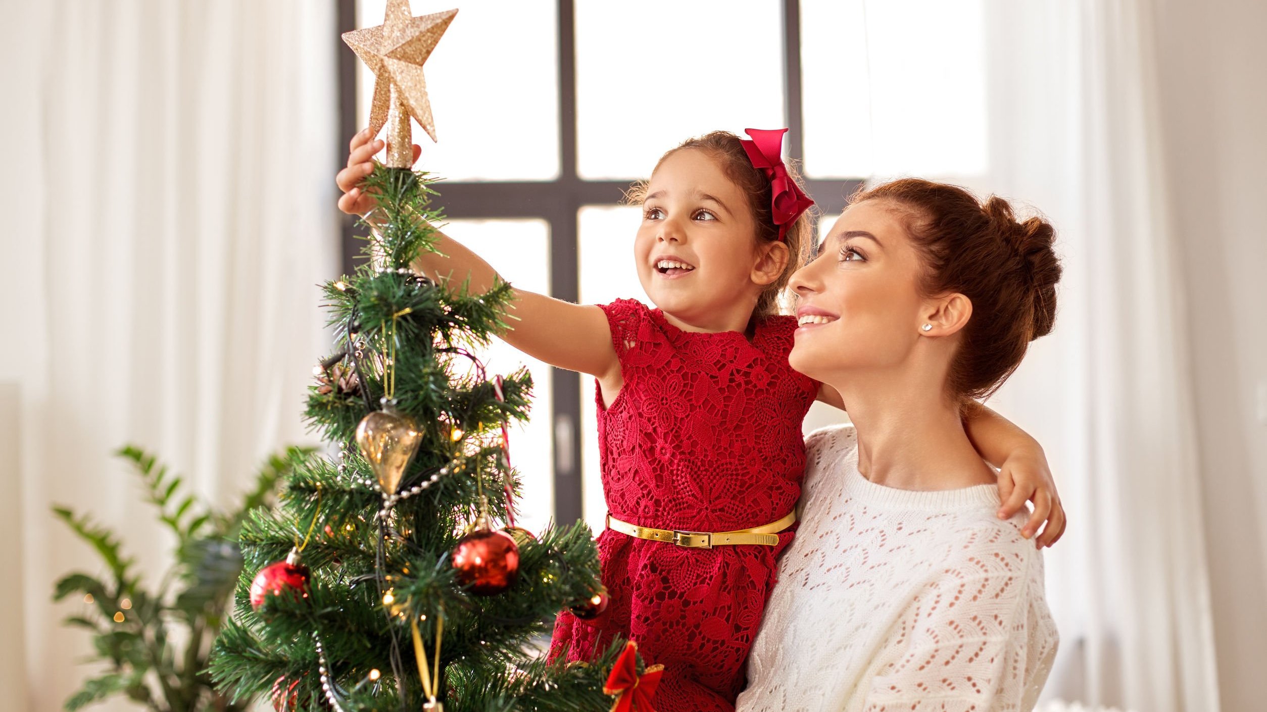dolgachov/ 123RF Mãe segurando filha no colo e filha colocando enfeite em árvore de natal