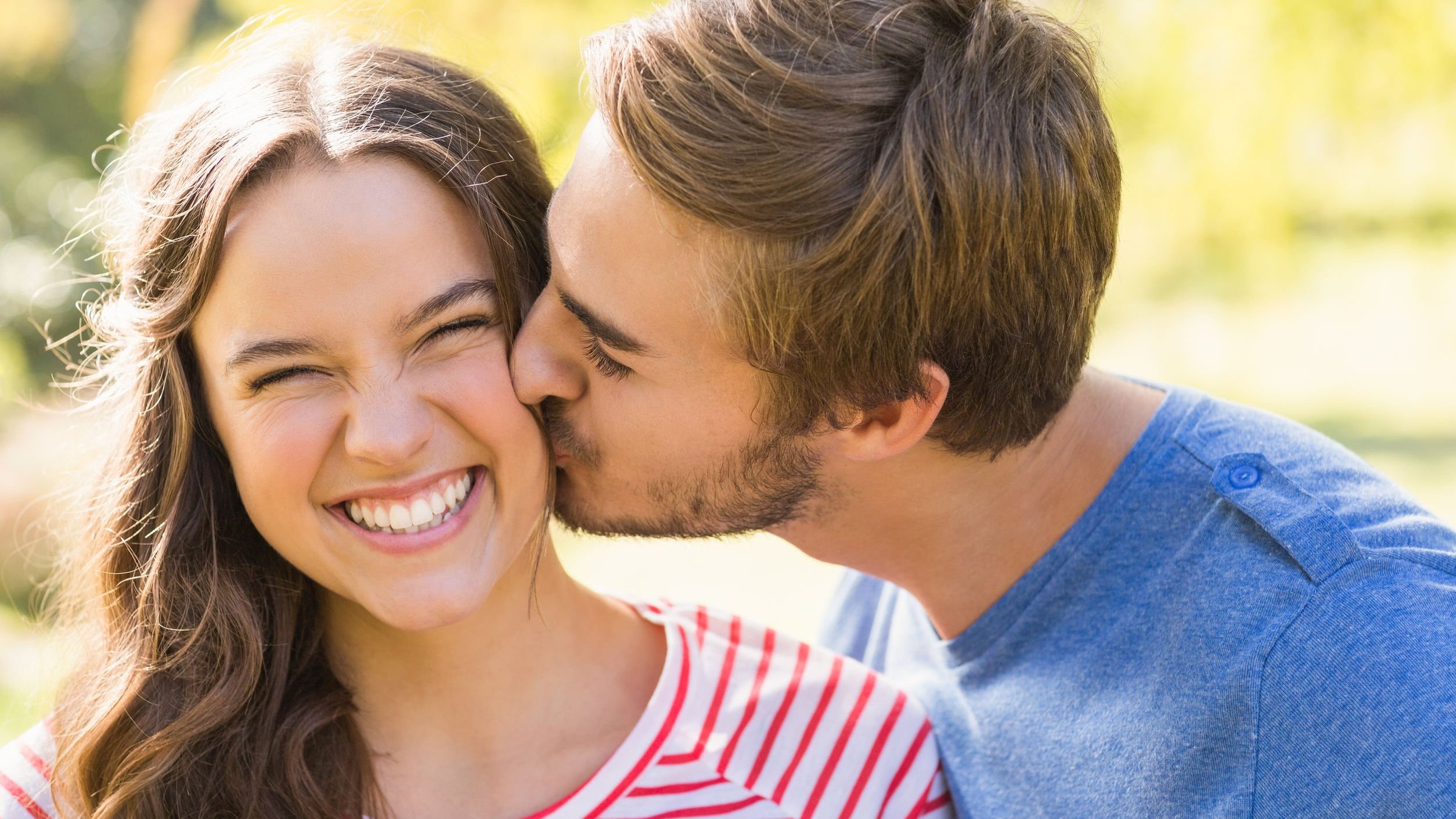 Homem beijando rosto de mulher sorrindo, ambos estão ao ar livre, durante um dia ensolarado