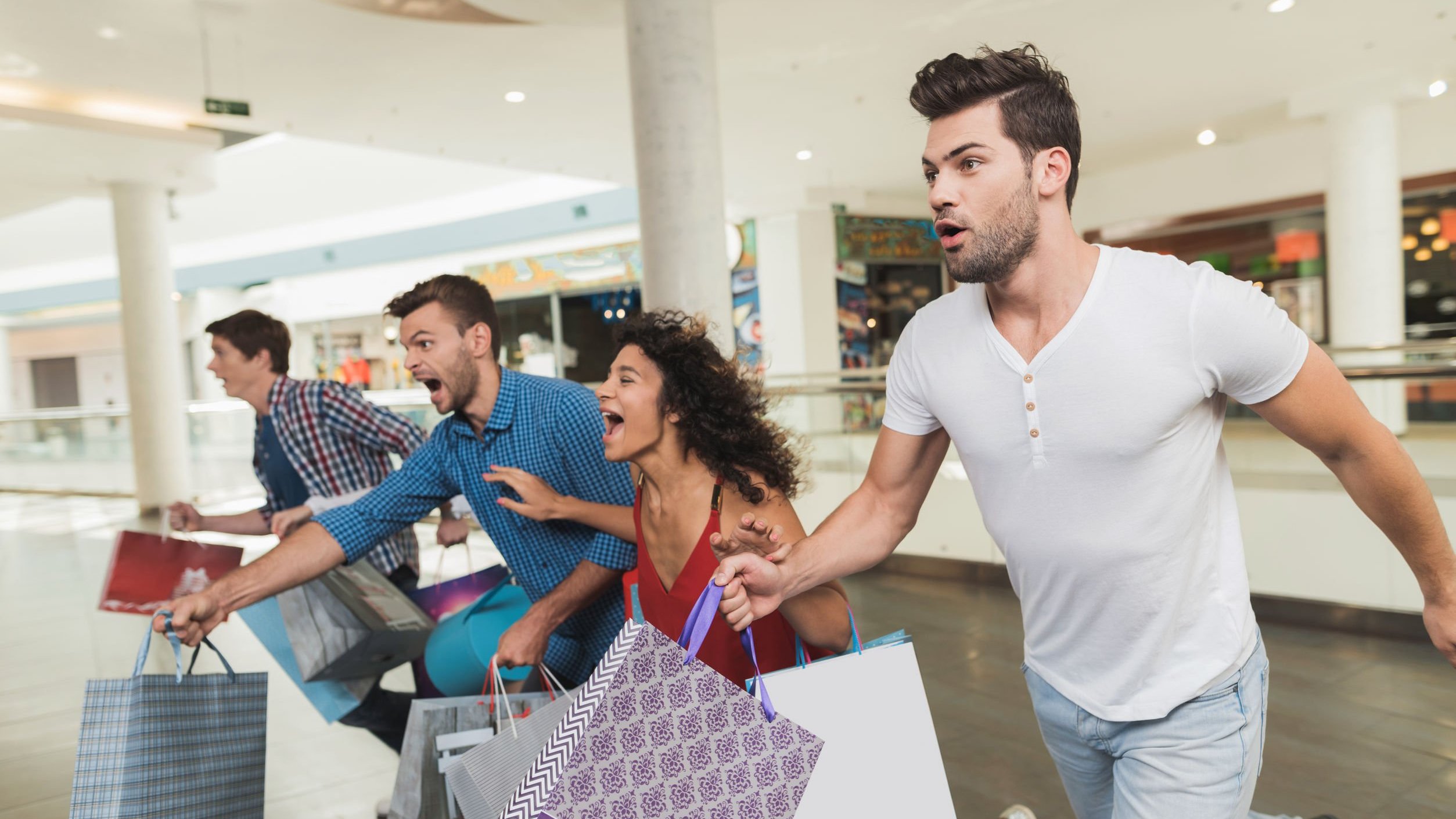 Quatro pessoas segurando sacolas de compras dentro de um shopping, com as bocas abertas como se estivessem gritando, e correndo.