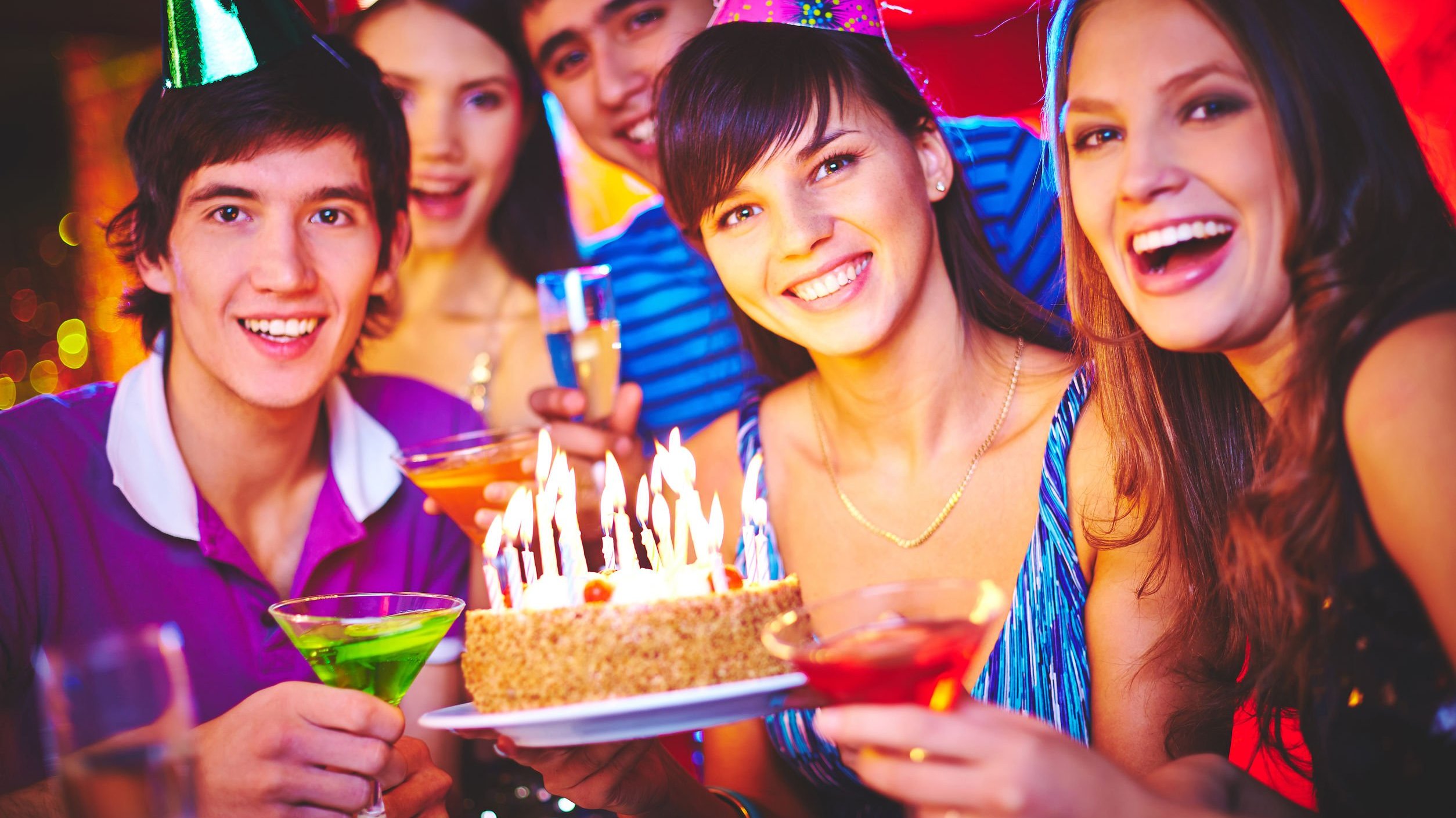 Pessoas comemorando aniversário ao redor de um bolo com velas acesas, todos sorrindo e usando chapéus de aniversário