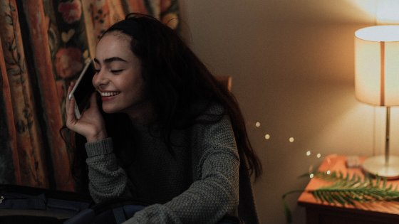 Menina conversando no telefone e sorrindo em seu quarto ao lado de uma cômoda com abajur